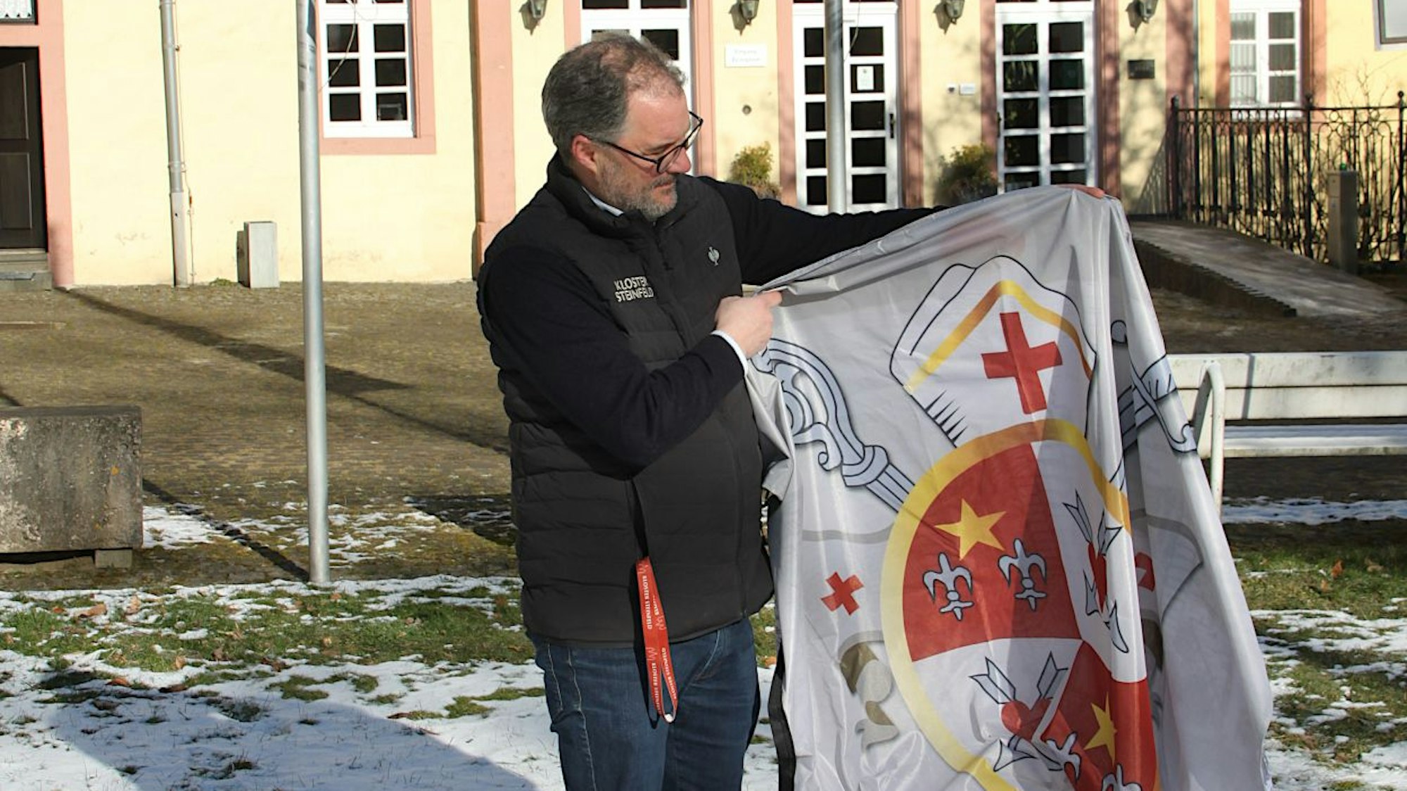 Christoph Böhnke steht in einem Innnenhof. Er hält eine neue Fahne des Klosters Steinfeld in den Händen und betrachtet sie.
