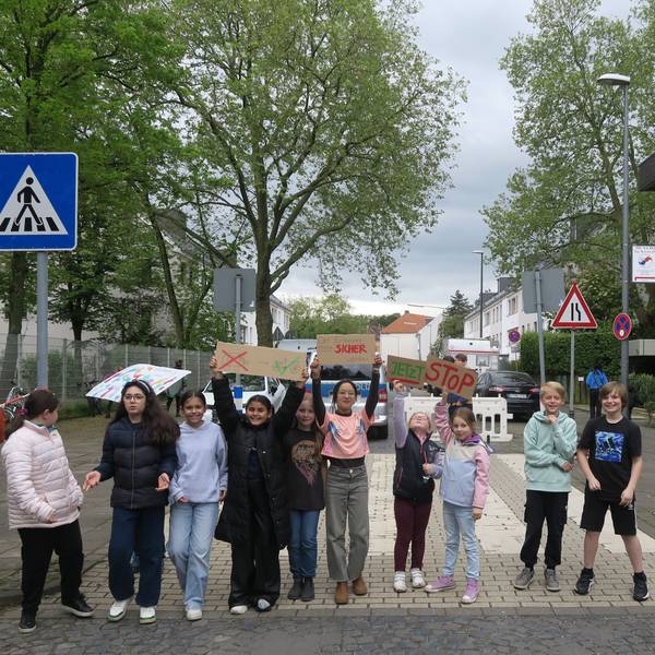 Mehrere Kinder stehen in einer Reihe und blockieren so eine Straße. Sie halten Plakate hoch, auf denen sie einen sicheren Schulweg fordern.