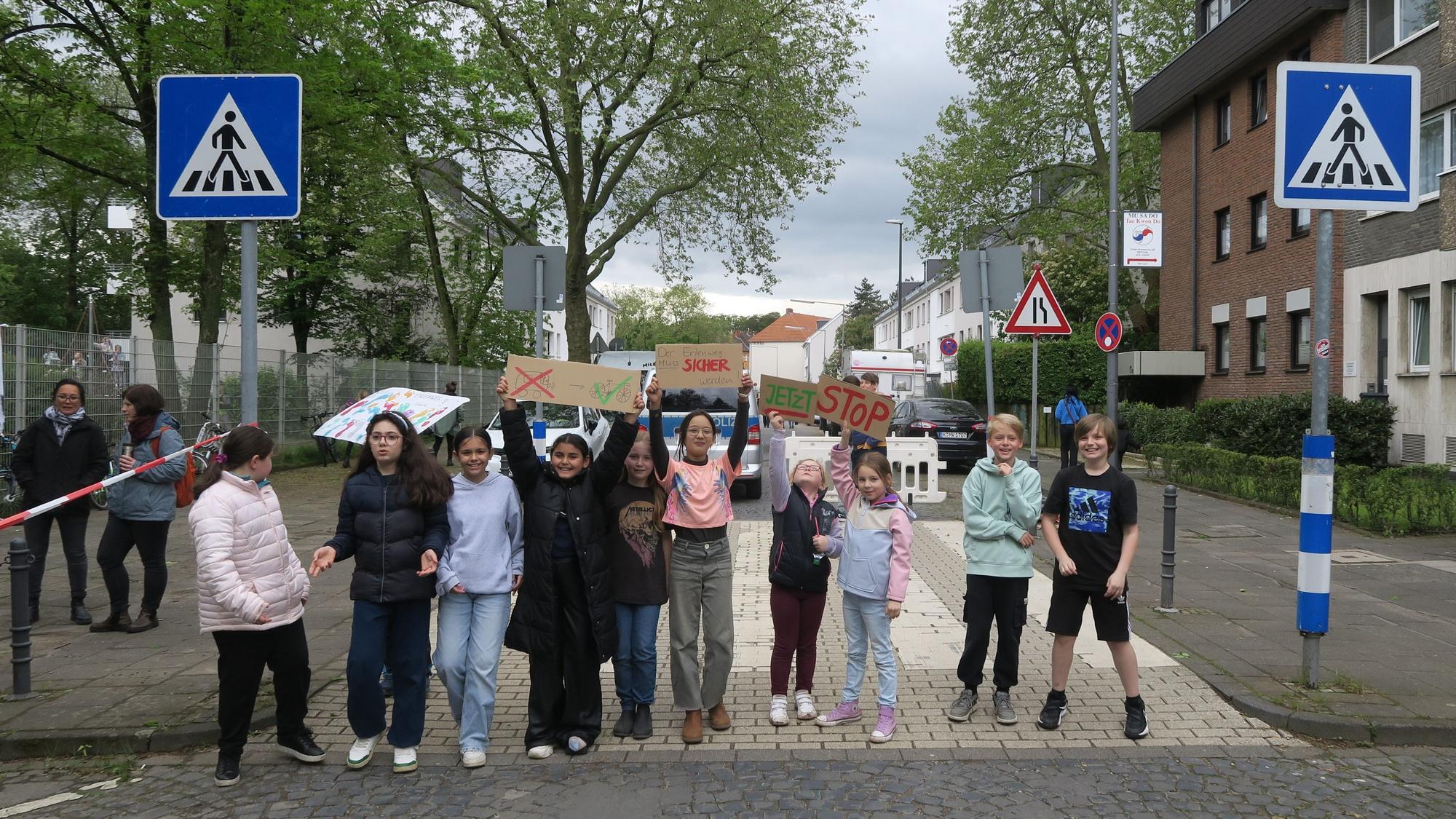 Mehrere Kinder stehen in einer Reihe und blockieren so eine Straße. Sie halten Plakate hoch, auf denen sie einen sicheren Schulweg fordern.