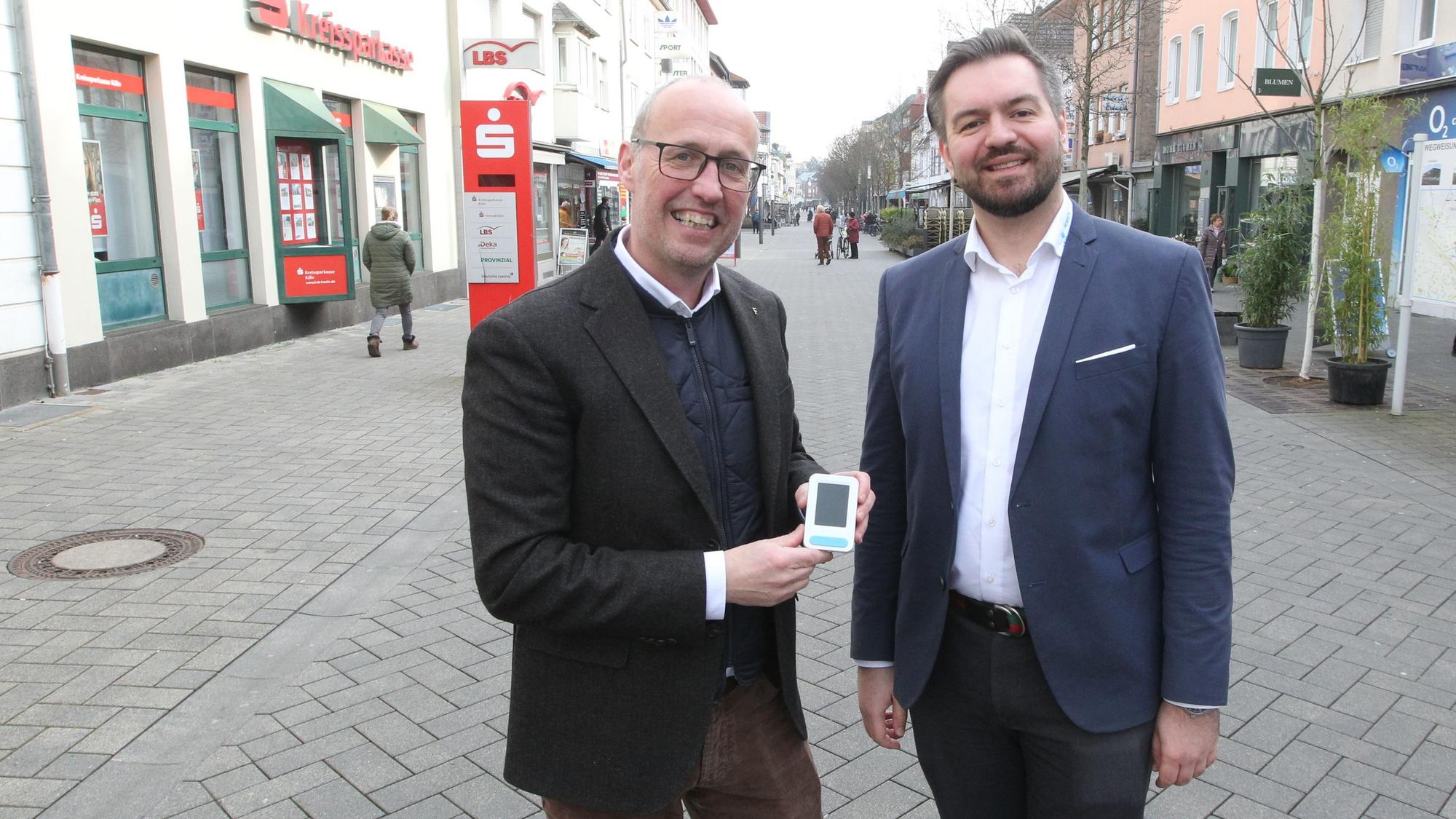 Zwei Männer stehen in einer Fußgängerzone. Einer zeigt ein kleines elektronisches Gerät.