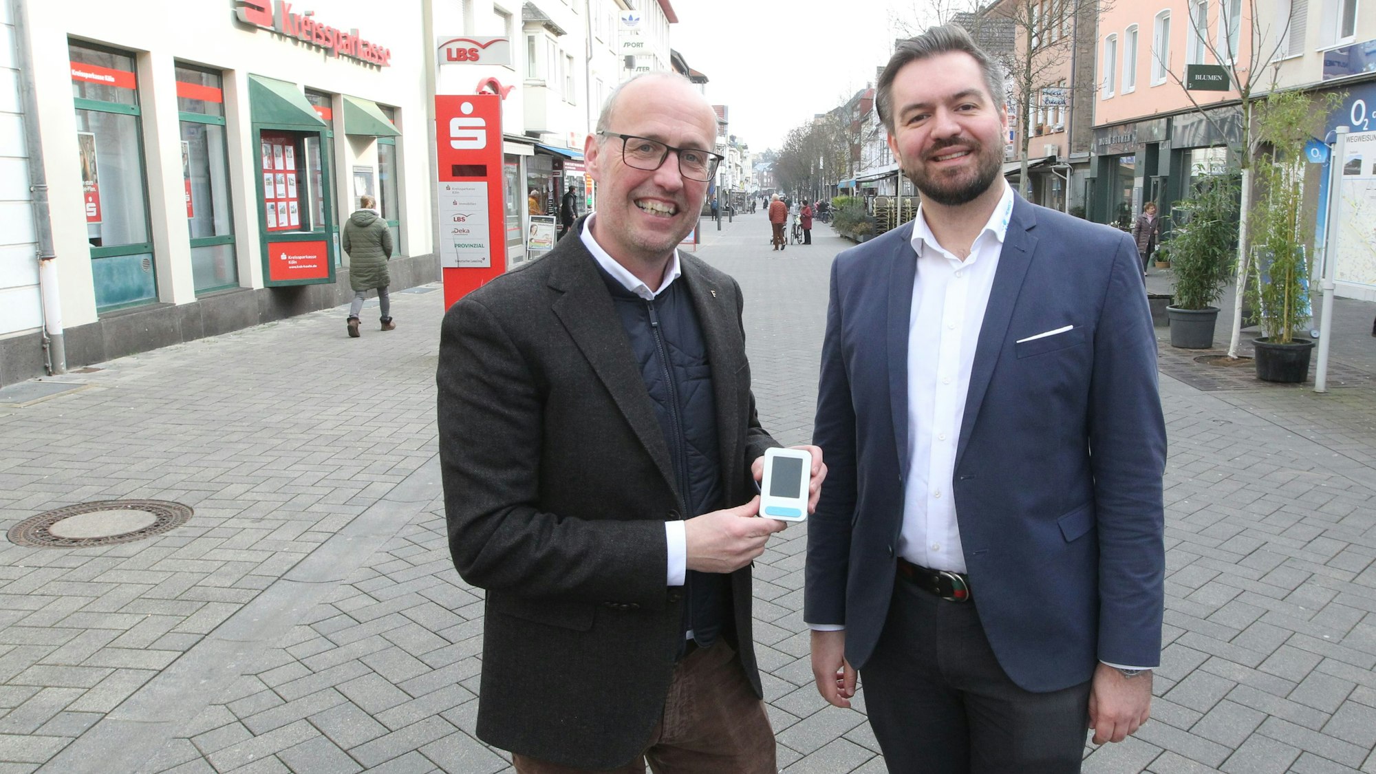 Zwei Männer stehen in einer Fußgängerzone. Einer zeigt ein kleines elektronisches Gerät.
