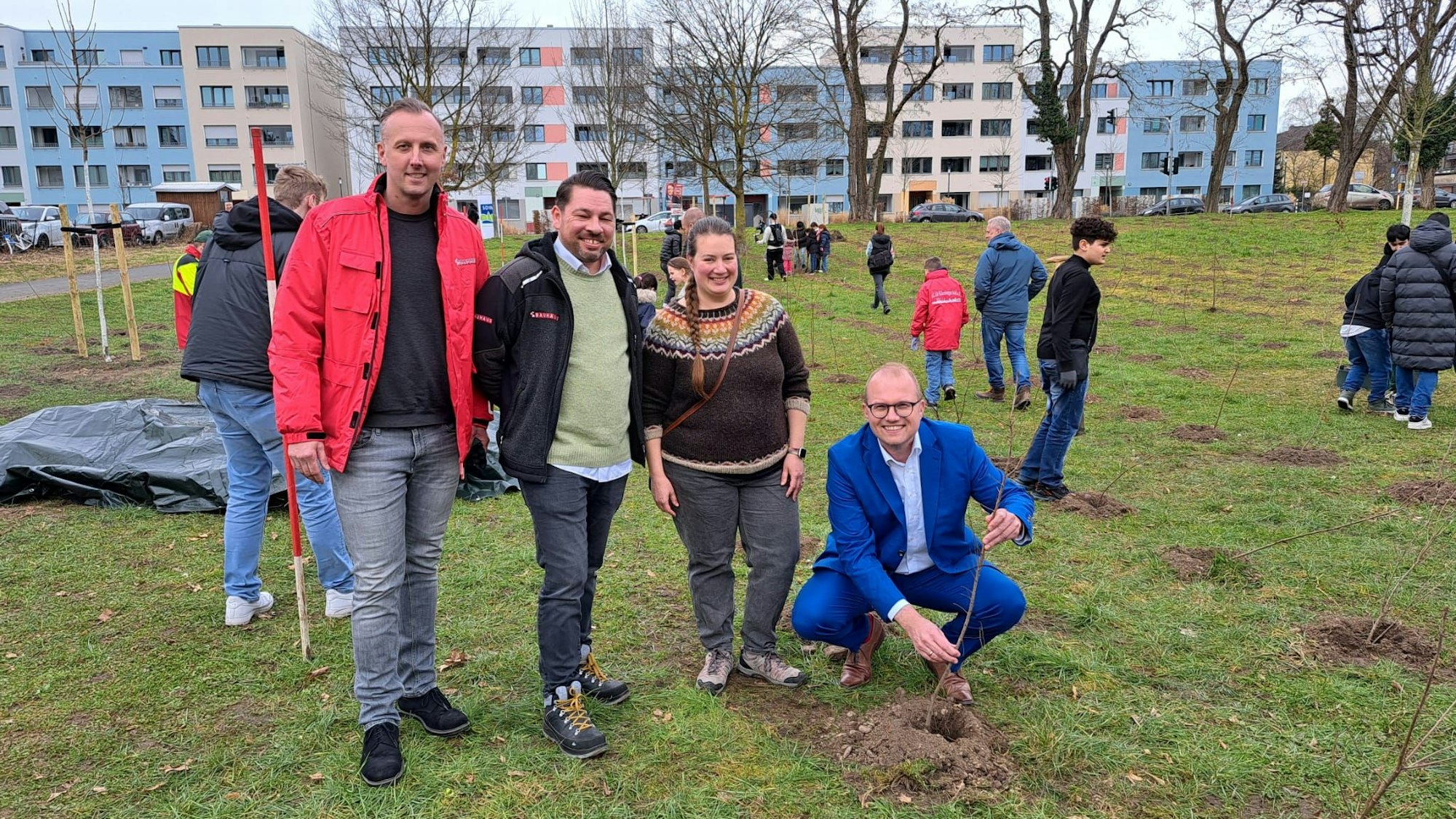 Drei Männer und eine Frau stehen und hocken auf einer Wiese, um sie herum sind viele Kinder. Ein Mann hält einen jungen Baum, der gerade gepflanzt wurde.