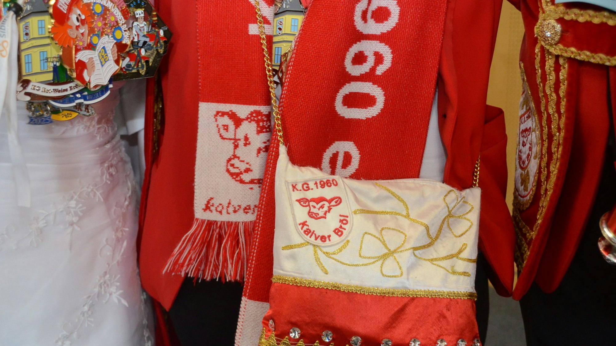 Eine kleine Umhängetasche aus glänzendem Stoff in Rot und Weiß. Auf dem Wappen ist eine Kuh zu sehen, außerdem die Aufschrift „KG Kalver Bröl.“