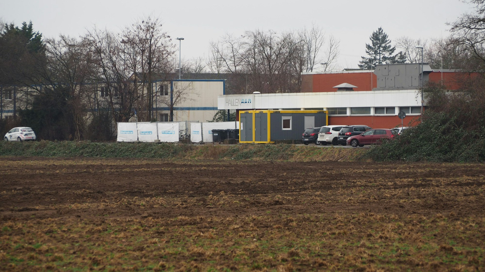 Ein Acker, dahinter das alte Hallenbad und Unterkünfte für Geflüchtete.