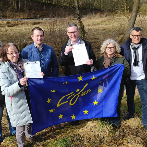 Die Genannten stehen vor einer Brachfläche an der Urft. Sie halten eine EU-Fahne und Vertragsunterlagen hoch.