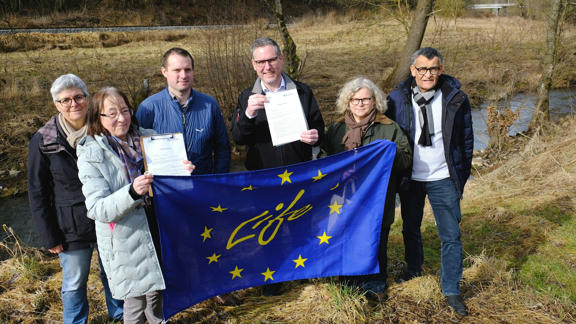 Die Genannten stehen vor einer Brachfläche an der Urft. Sie halten eine EU-Fahne und Vertragsunterlagen hoch.