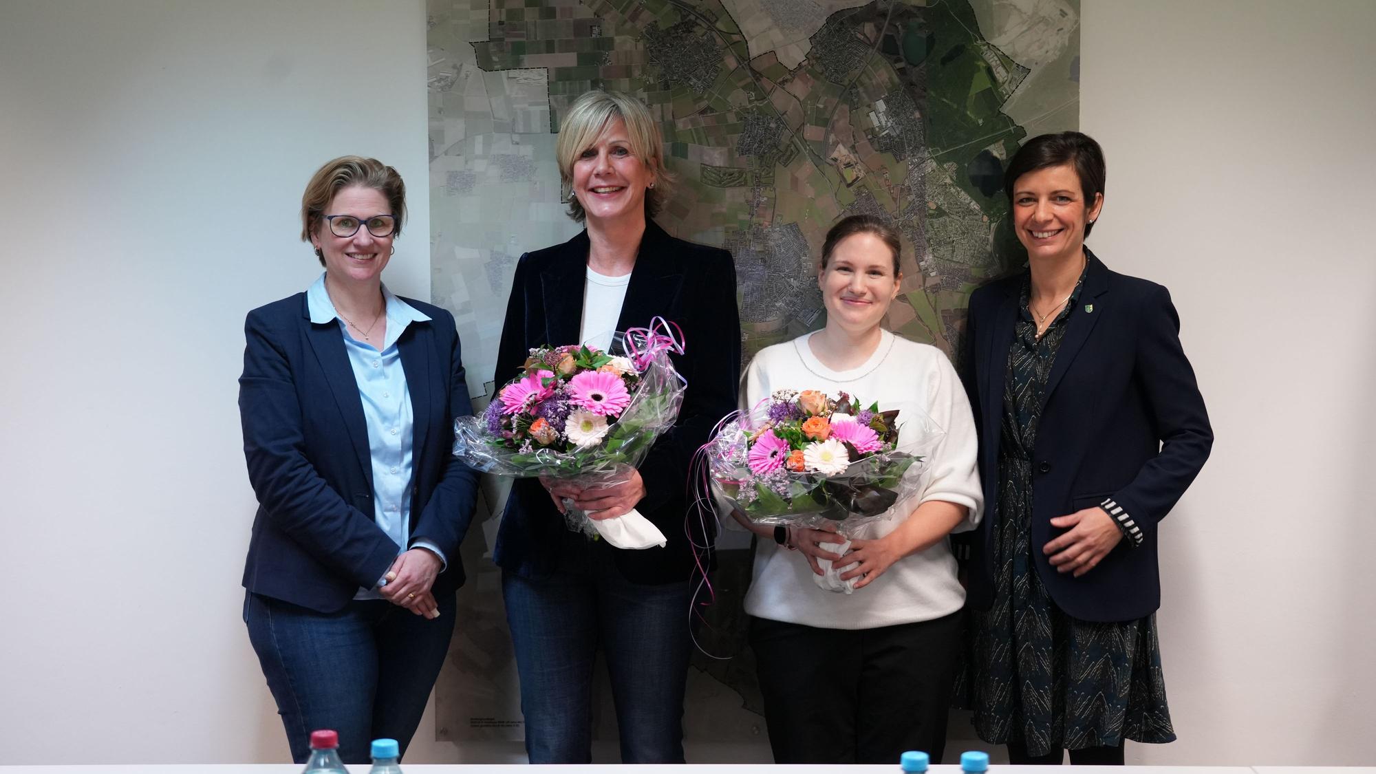 Vier Frauen lächeln in die Kamera, die beiden in der Mitte halten Blumensträuße in ihren Händen.