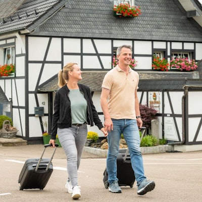 Zwei Menschen ziehen einen Rollkoffer hinter sich her. Im Hintergrund ist ein Fachwerkhaus zu sehen.