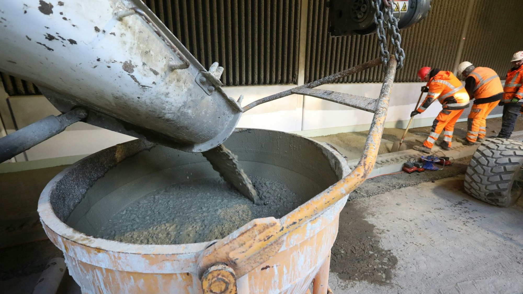 Beton fließt aus einem Betonmischer in einen hohen Behälter, im Hintergrund verarbeiten Arbeiter den Beton.