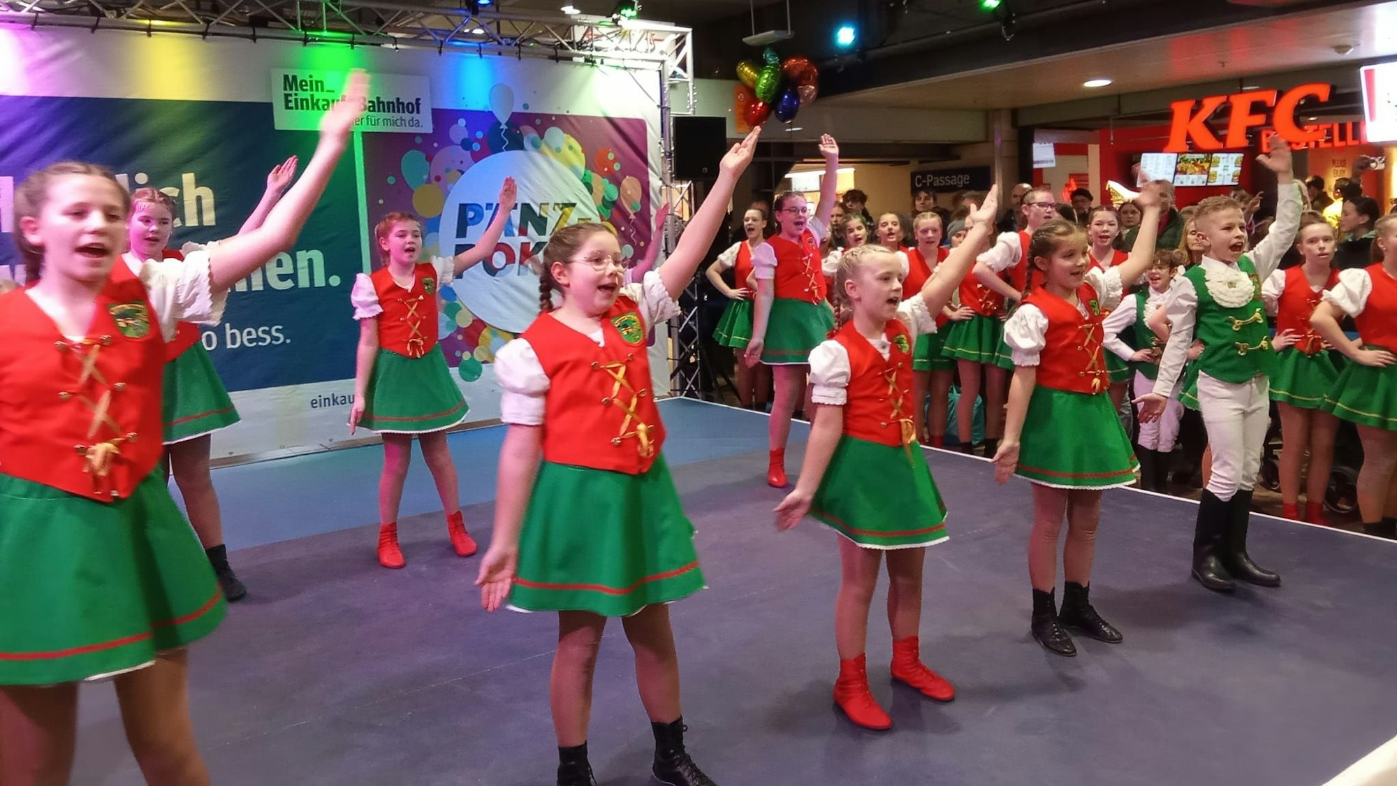 Die Kinder tragen rote Westen und grüne Röcke, während sie im Bahnhof ihren Tanz aufführen.