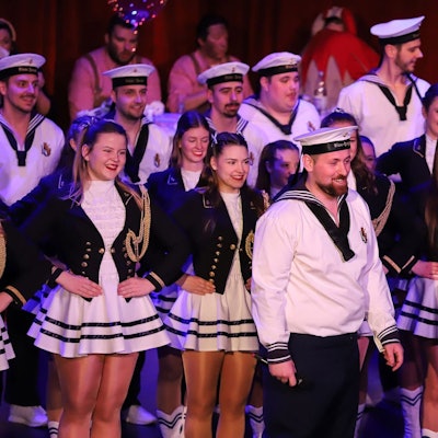 Auf dem Bild ist ein Mann im Matrosenkostüm vor einer Reihe Tänzer auf der Bühne zu sehen.
