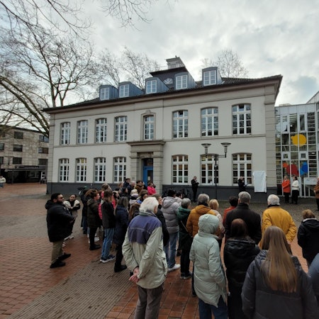 Auf dem Bild ist die verhüllte Infotafel an der VHS sowie Publikum zu sehen.