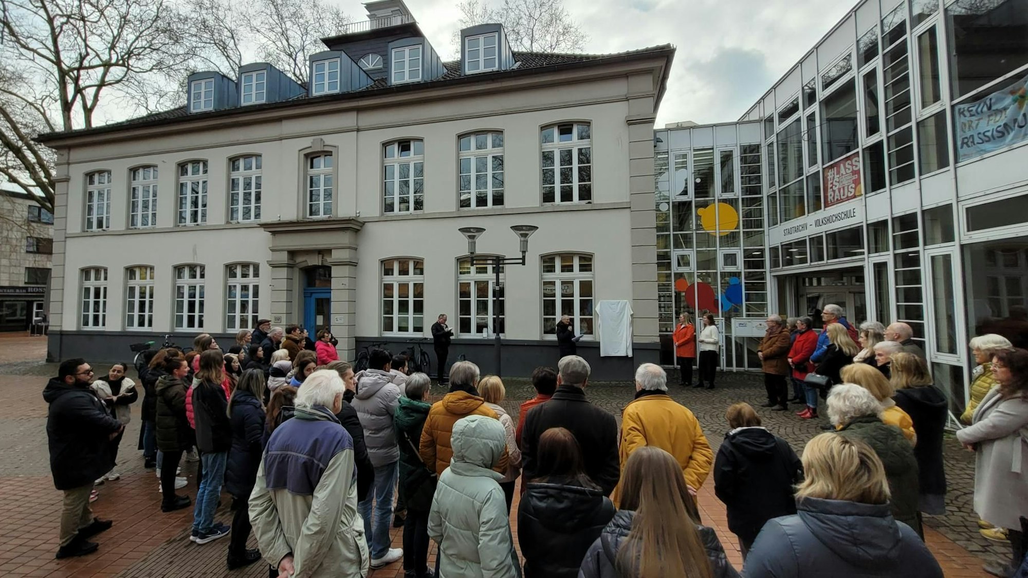 Auf dem Bild ist die verhüllte Infotafel an der VHS sowie Publikum zu sehen.