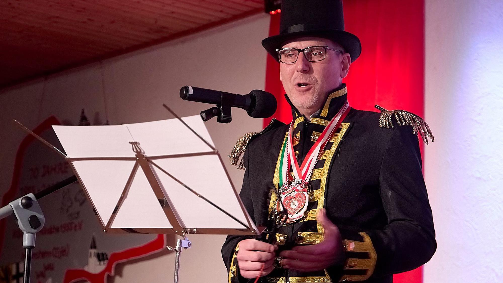 Nettersheims Bürgermeister Norbert Crump steht als Dompteur verkleidet an einem Notenständer, auf dem zwei Blätter sind. Er spricht in ein Mikrofon.