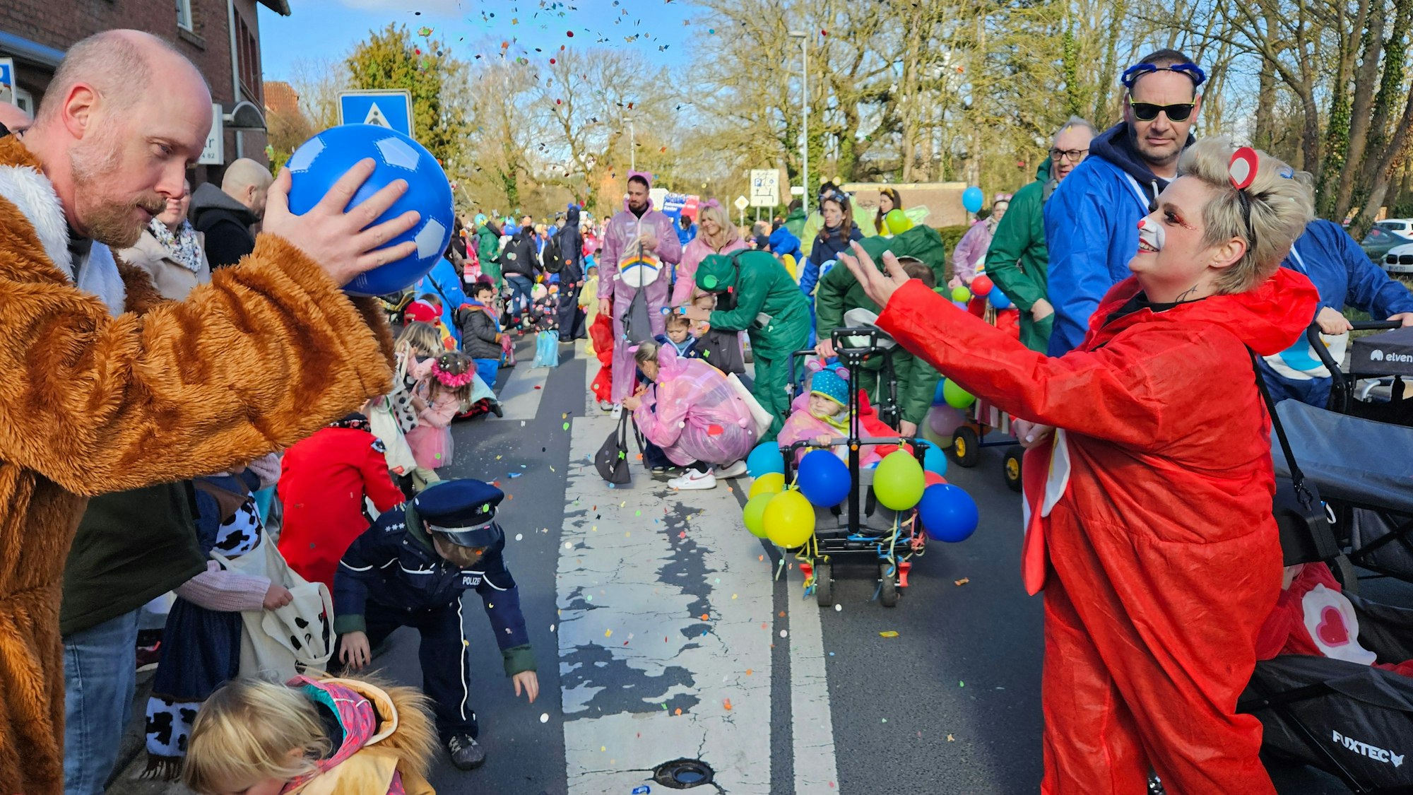 Zu sehen sind zahlreiche kostümierte Menschen in einem Karnevalszug, eine Frau wirft Konfettig, ein Mann fängt einen Ball.