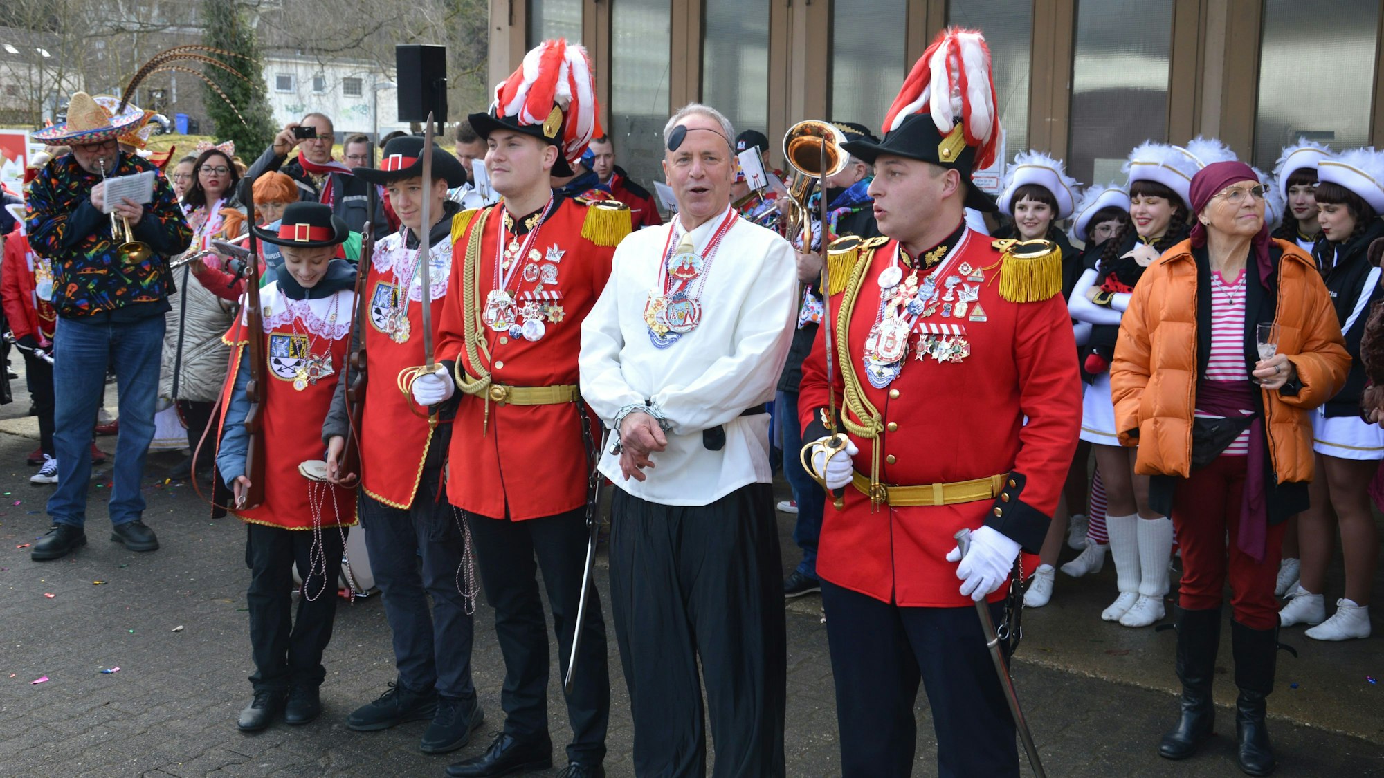 Kalls Bürgermeister Hermann-Josef Esser trägt ein weißes Hemd und schwarze Hose, seine Augenklappe hat er auf die Stirn geschoben. Er steht in Handschellen inmitten kostümierter Karnevalisten.