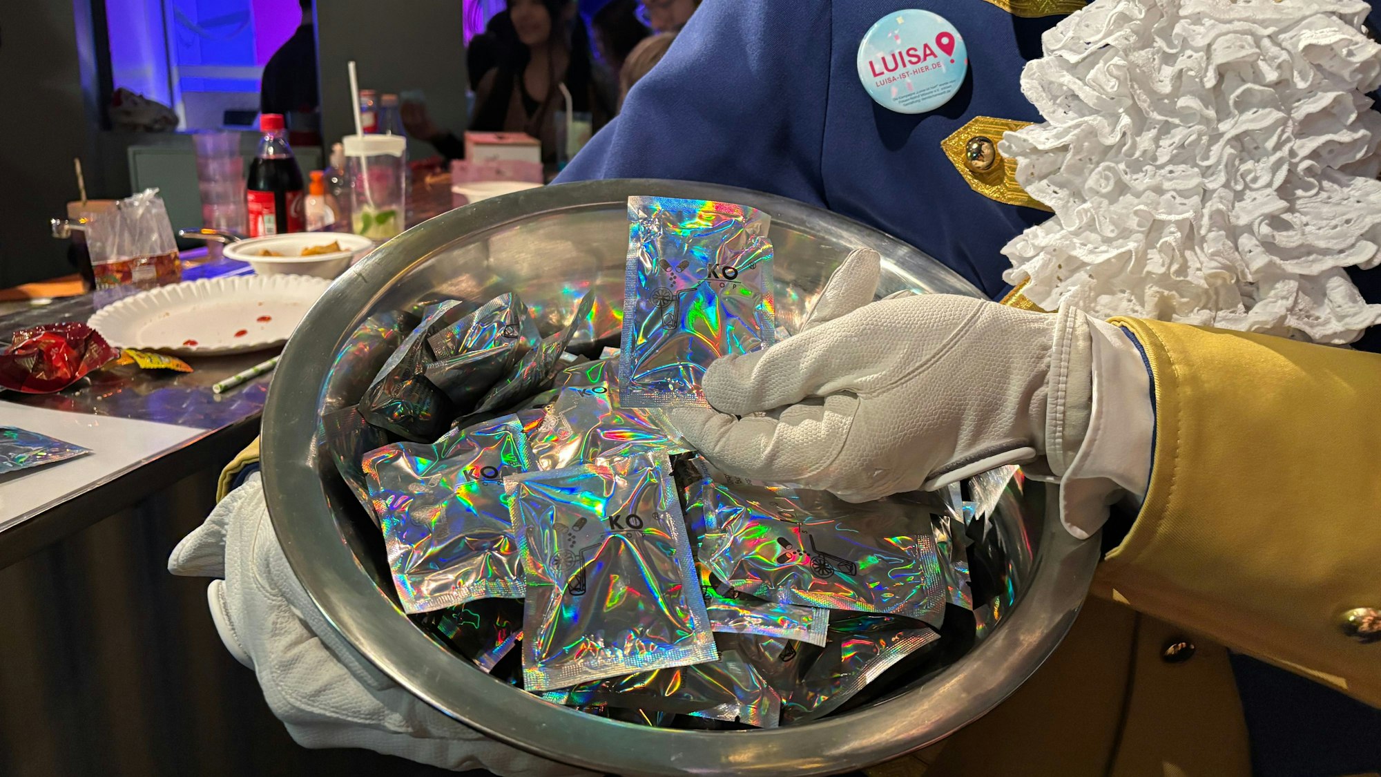In einer Schüssel liegen kleine Verpackungen, die das Licht, was auf sie fällt, regenbogenfarben reflektieren. Eine Hand in Handschuh hält eine der Verpackungen ins Bild.