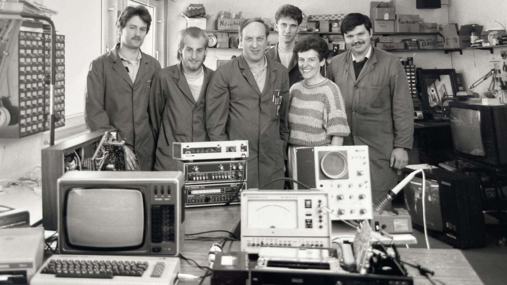 Männer in Arbeitskitteln und eine Frau im gestreiften Pullover posieren auf einem Schwarz-Weiß-Foto hinter Elektrogeräten auf einem Tisch.