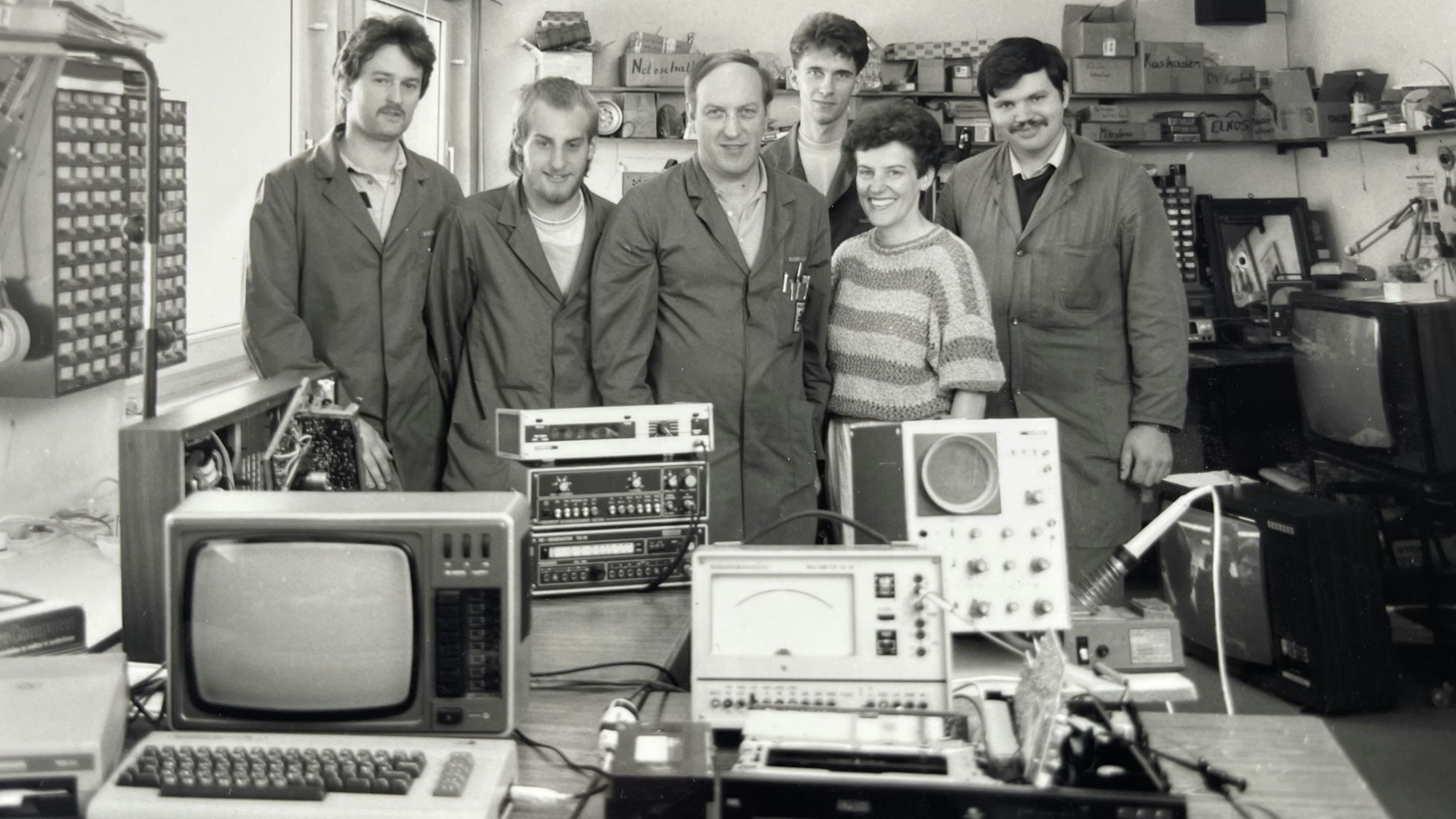 Männer in Arbeitskitteln und eine Frau im gestreiften Pullover posieren auf einem Schwarz-Weiß-Foto hinter Elektrogeräten auf einem Tisch.