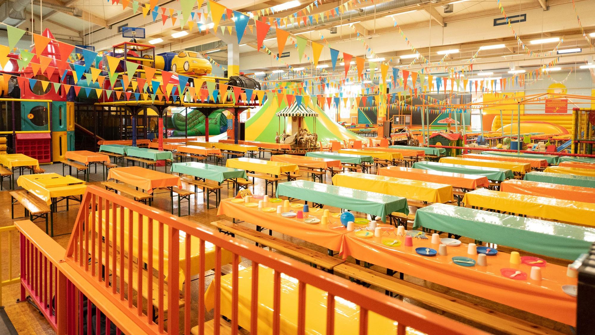 Sitzbänke in knalligen Farben stehen an ebenso bunten Tischen. Im Hintergrund befindet sich ein Indoorspielplatz.
