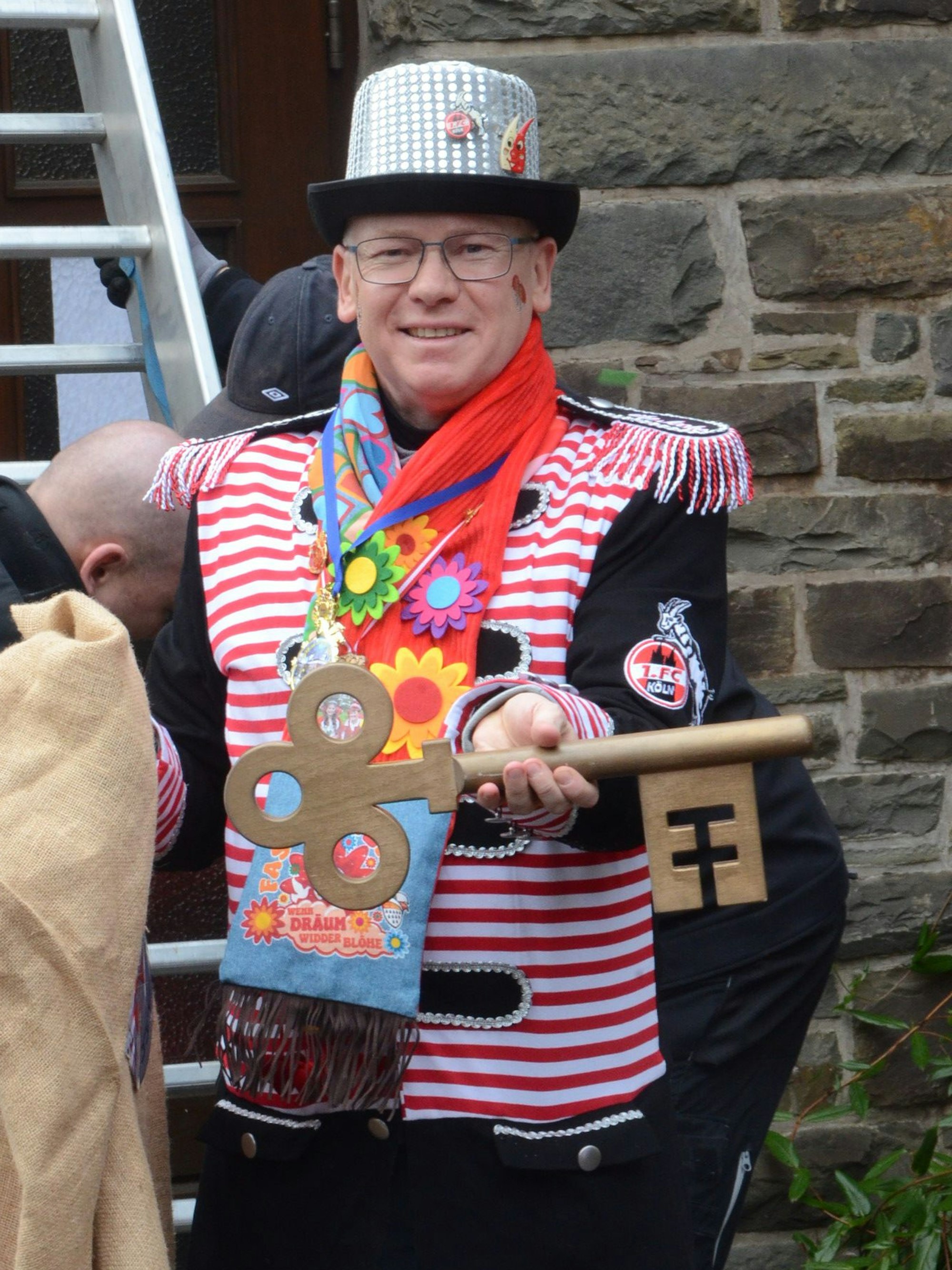 iEin bunt gekleideter Mann mit glitzerndem Zylinder und einem bunten Schal; in der Hand hält er einen großen nachgebildeten Schlüssel.