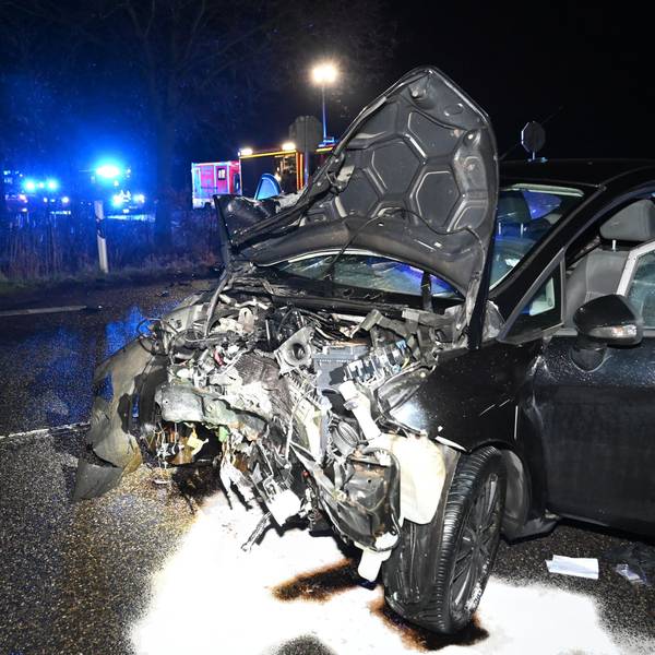 Ein dunkles Auto mit komplett zerstörter Motorhaube steht auf einer Straße. Im Hintergrund ist Blaulicht zu sehen.