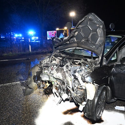 Ein dunkles Auto mit komplett zerstörter Motorhaube steht auf einer Straße. Im Hintergrund ist Blaulicht zu sehen.