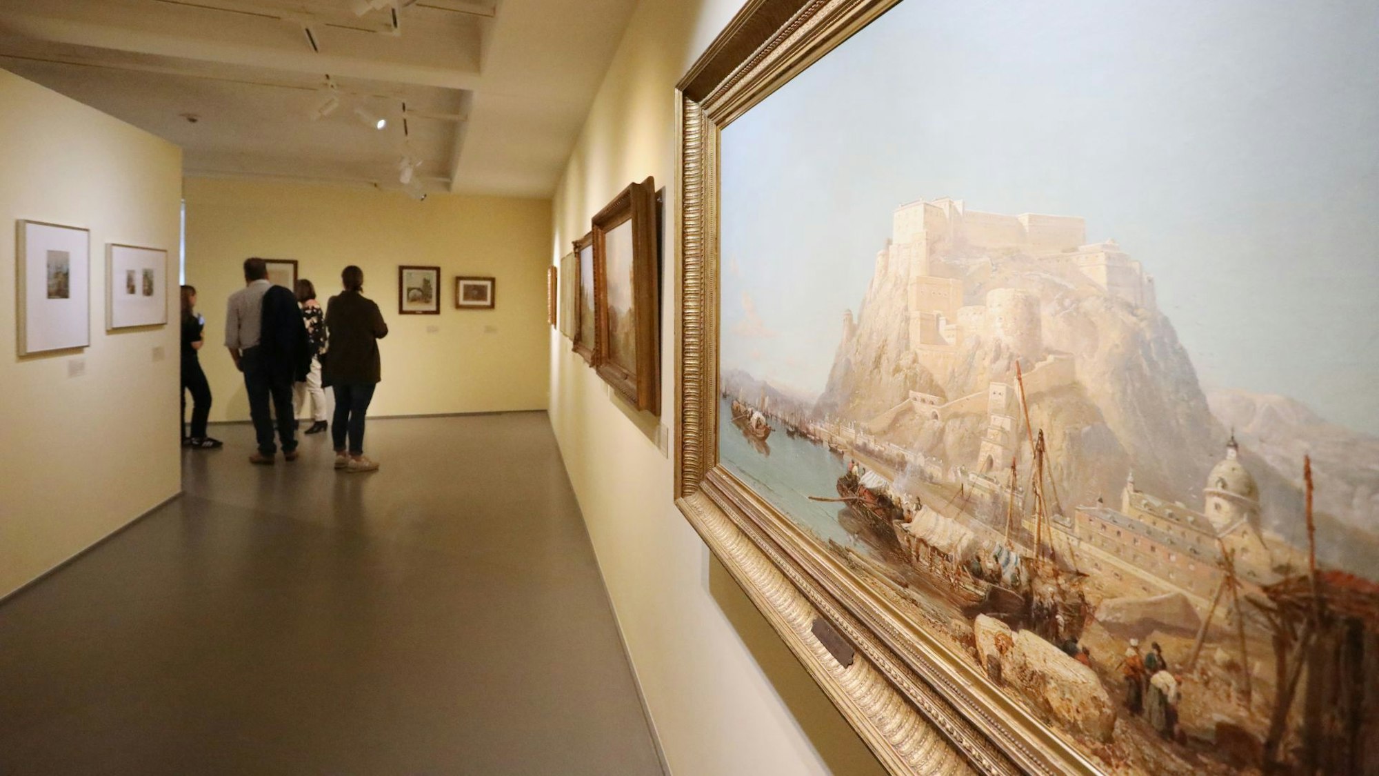 In der Aufnahme steht ein Gemälde an einer Wand im Fokus, auf dem Schiffe an einem Fluss und eine Berglandschaft abgebildet sind. Es sind zudem Museumsgäste zu sehen, die auf weitere Bilder schauen.