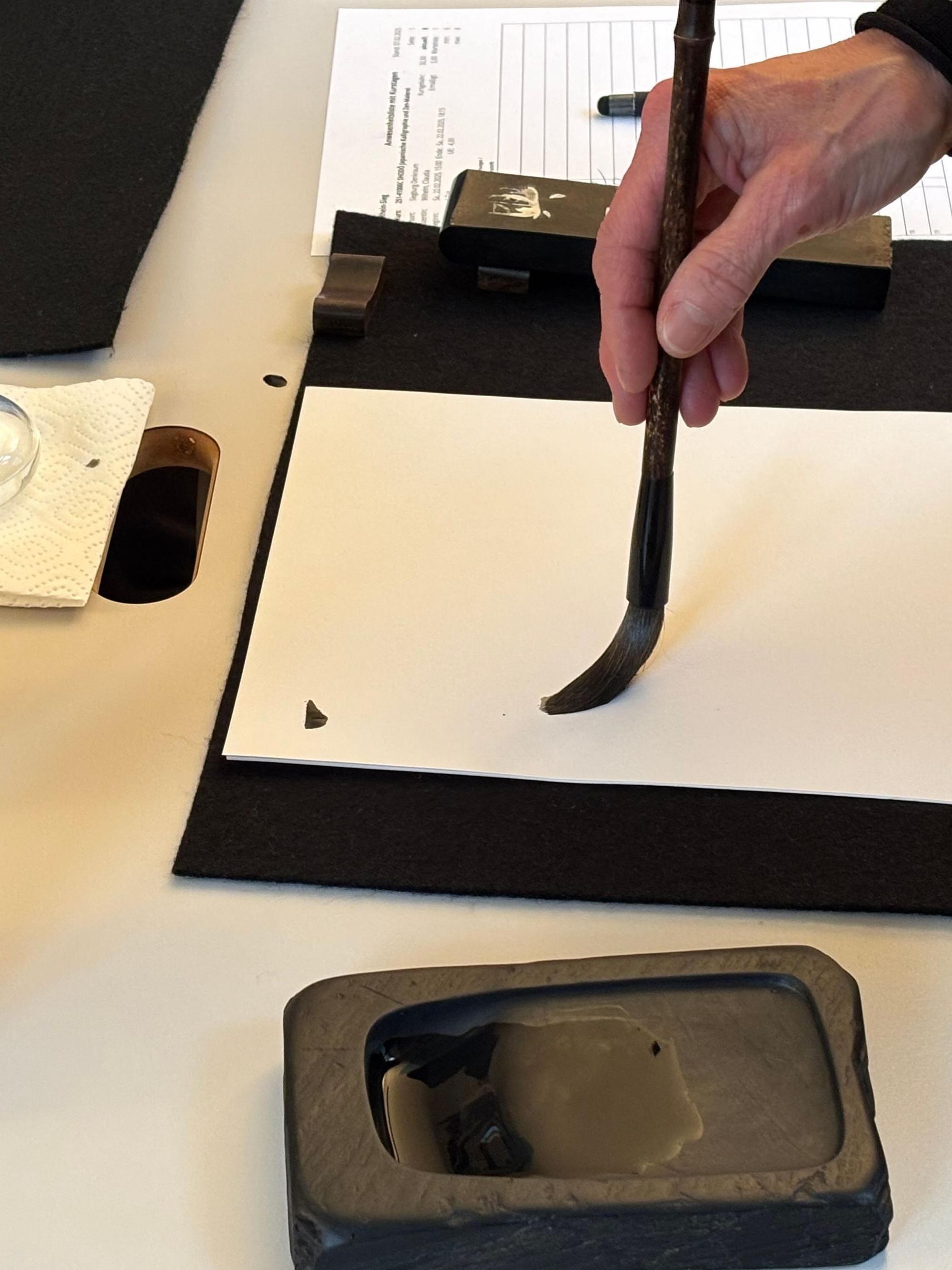 Nahaufnahme einer Hand, die mit einem Pinsel, der in Tusche getränkt wurde, auf einem Blatt Papier beginnt mit einem Strich zu Schreiben