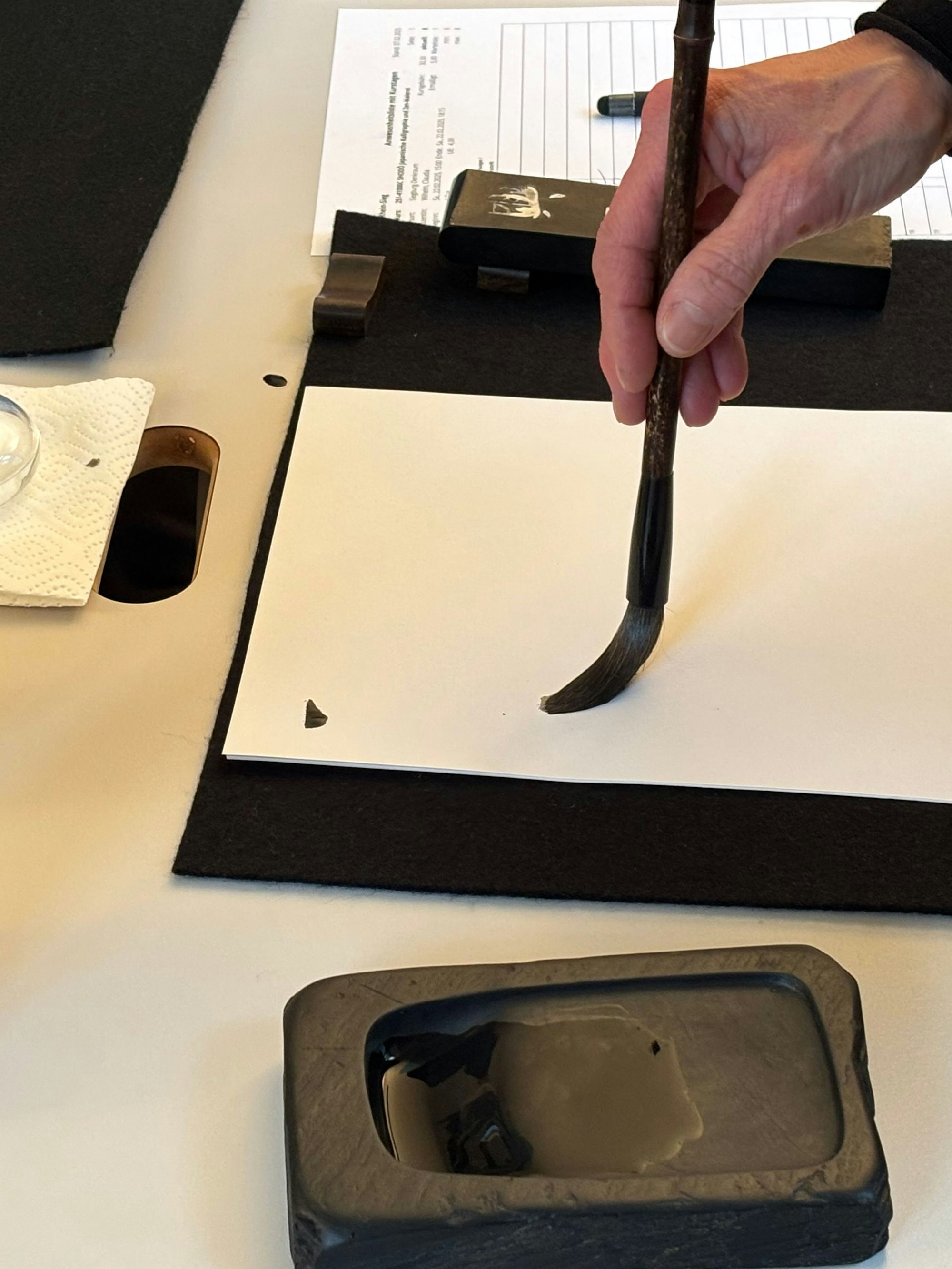Nahaufnahme einer Hand, die mit einem Pinsel, der in Tusche getränkt wurde, auf einem Blatt Papier beginnt mit einem Strich zu Schreiben