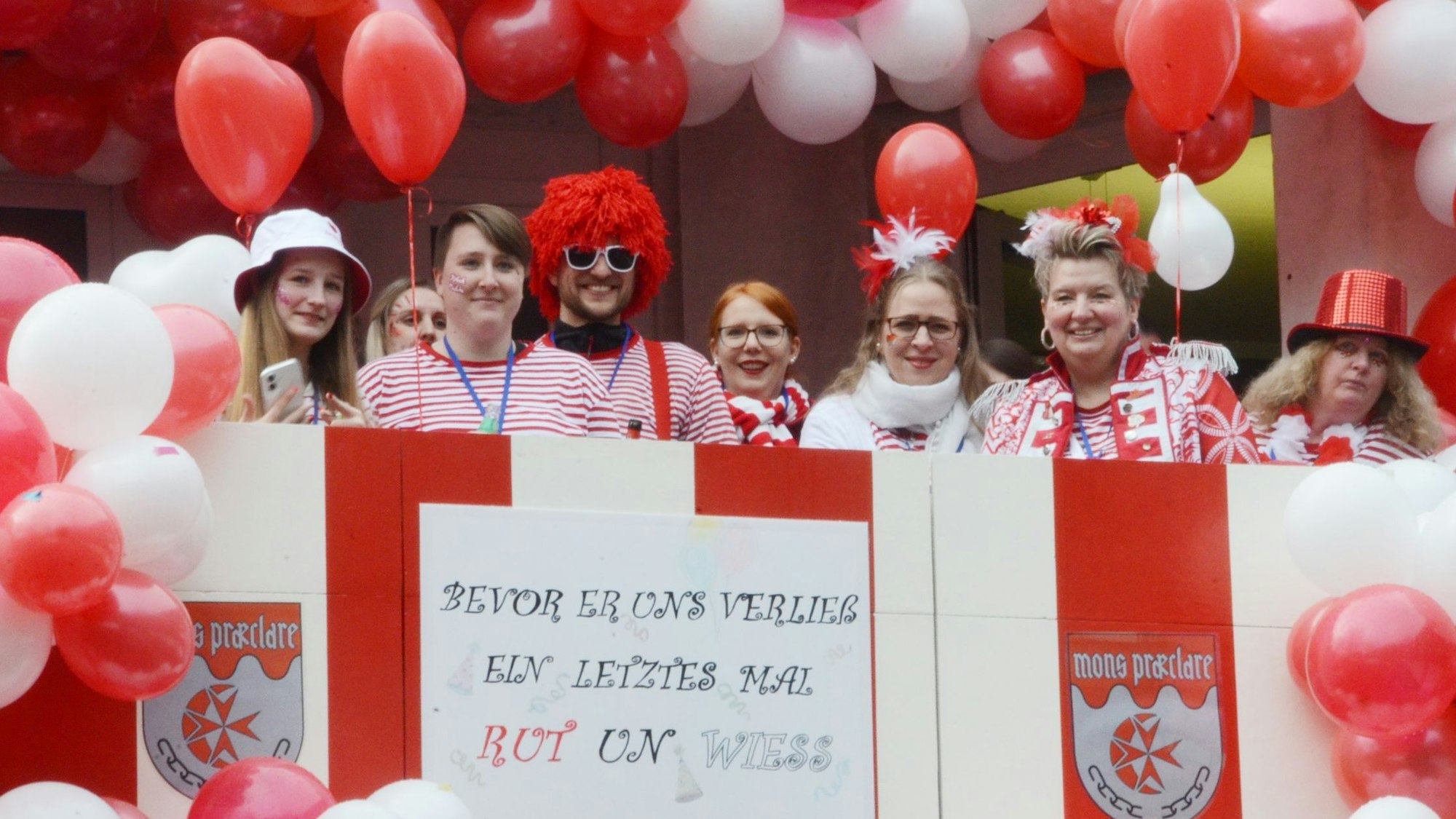 Männer und Frauen in rot-weißen Kostümen stehen auf einem ebenfalls in Rot und Weiß geschmückten Balkon.