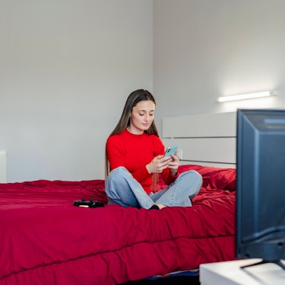 Eine junge Erwachsene sitzt auf ihrem Bett und schaut auf ihr Smartphone.