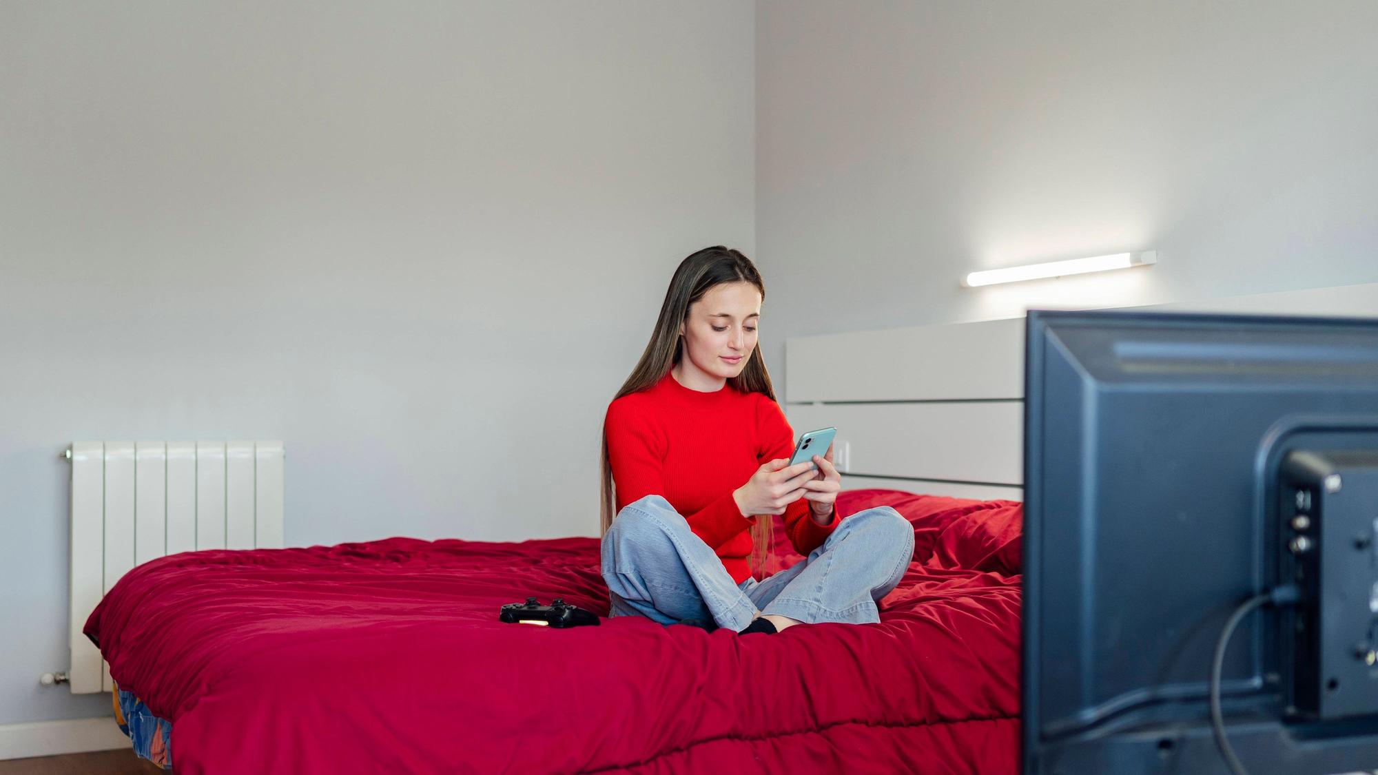 Eine junge Erwachsene sitzt auf ihrem Bett und schaut auf ihr Smartphone.