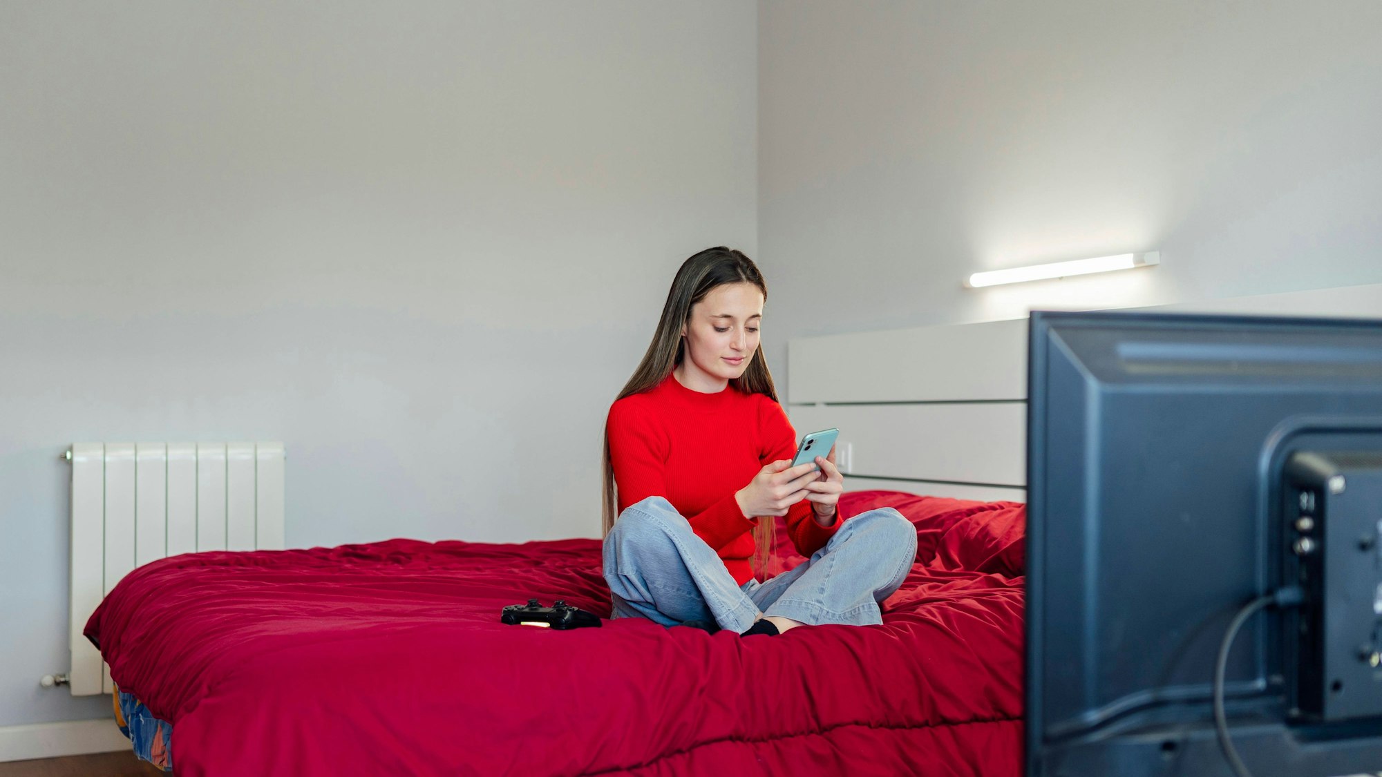 Eine junge Erwachsene sitzt auf ihrem Bett und schaut auf ihr Smartphone.
