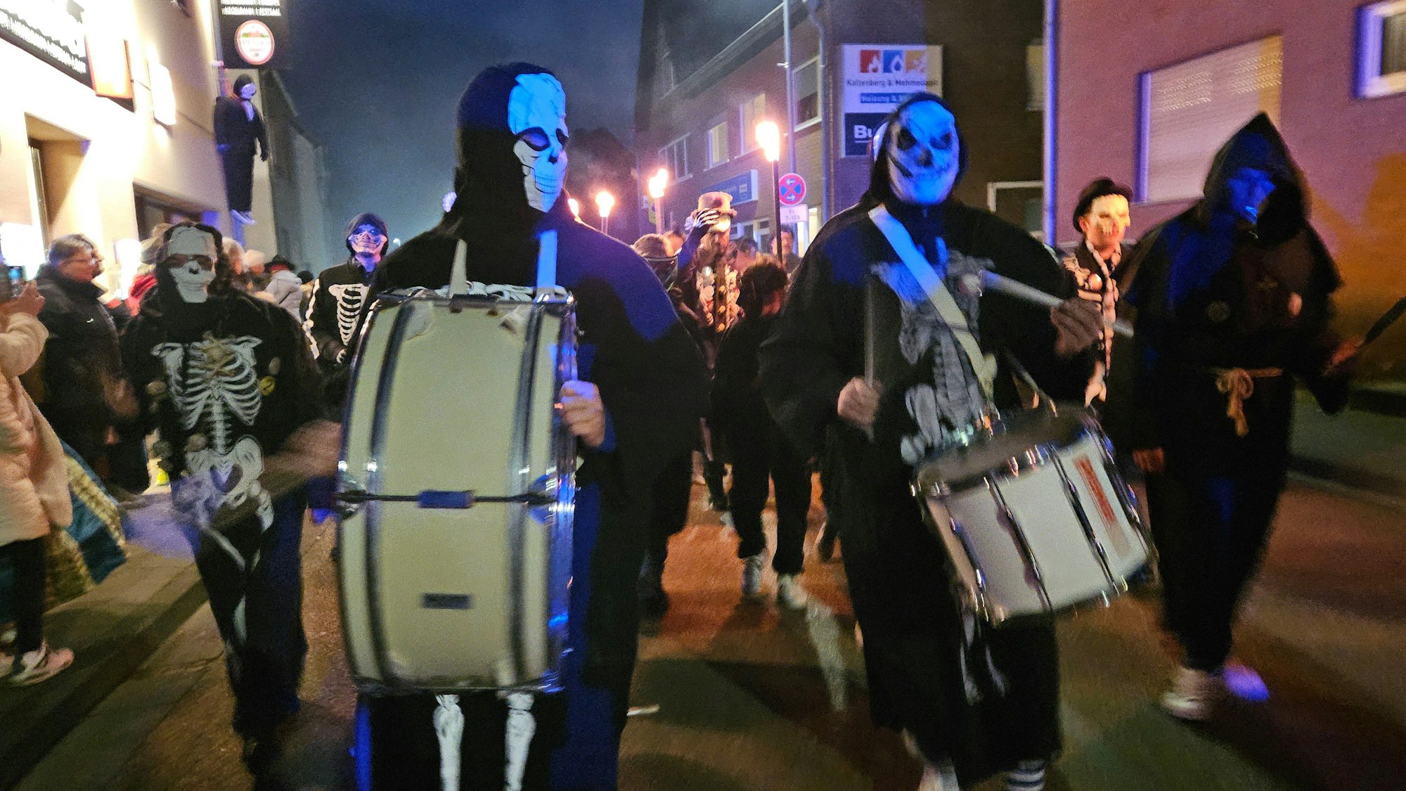 Zu sehen ist, wie Menschen, die als Geister kostümiert sind und Trommeln tragen, durch eine nächtliche Straße ziehen.