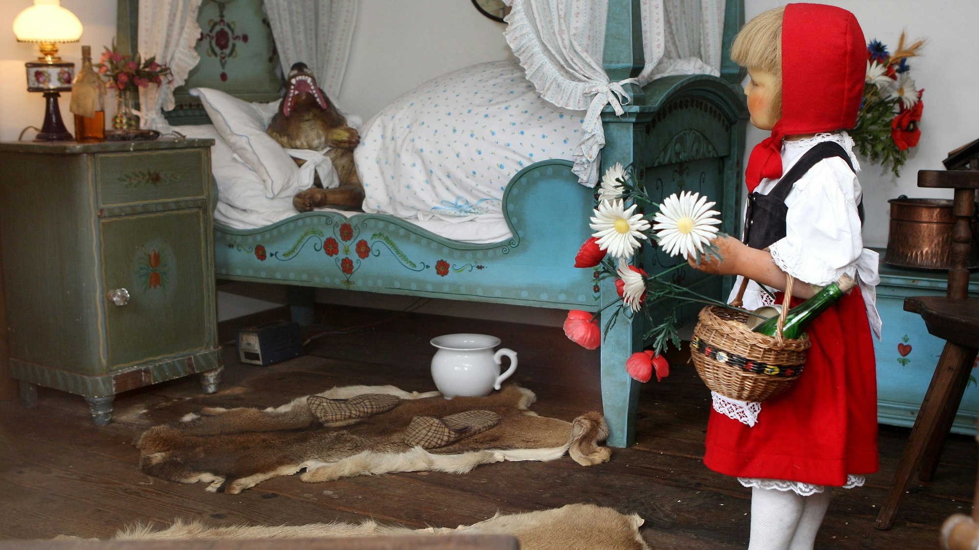 Eine Rotkäppchen-Figur steht mit einem Korb in einem Zimmer, in dem eine Wolf-Figur in einem Bett schläft.