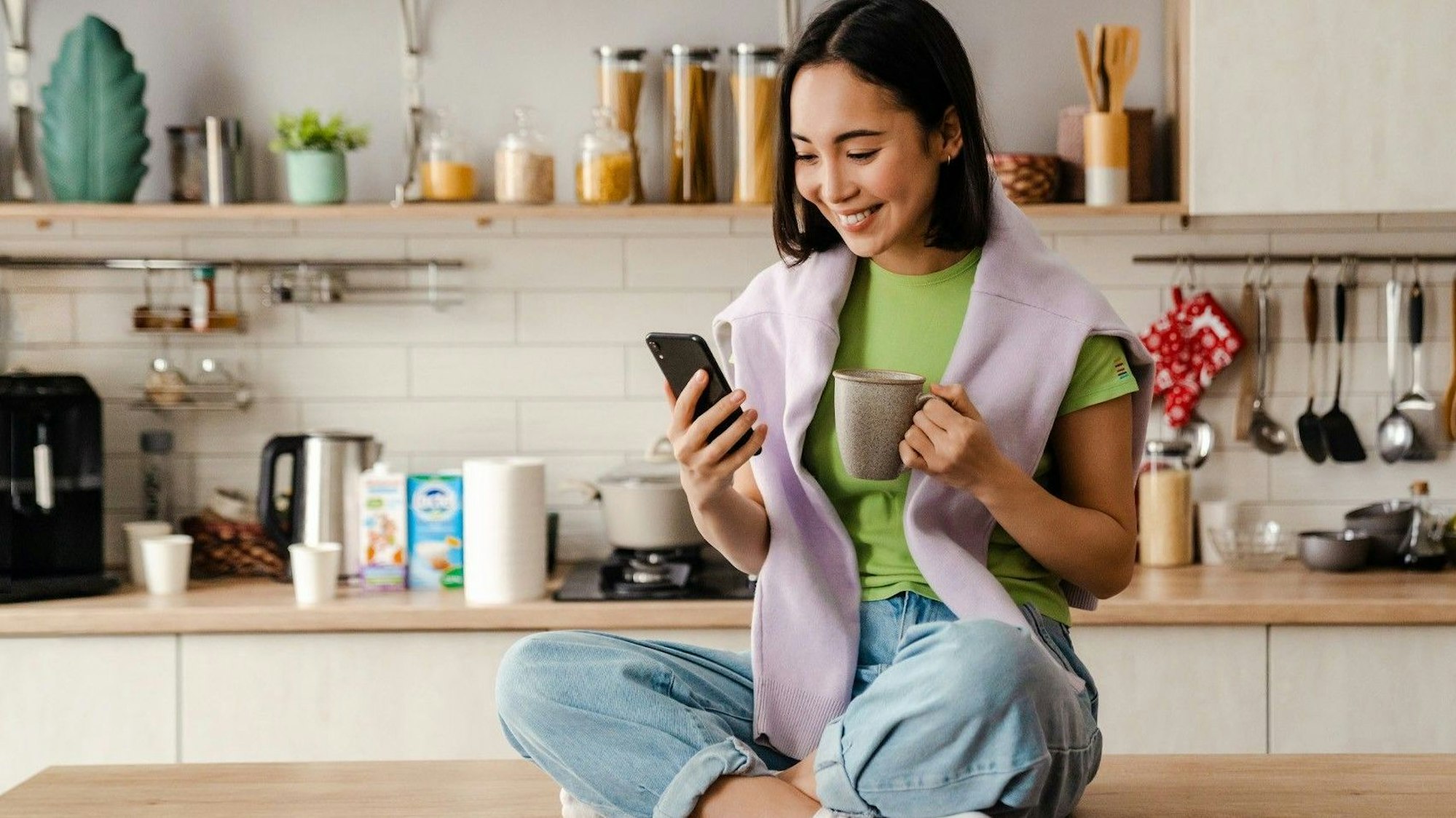Frau sitzt auf Küchentisch und schaut freudig auf Smartphone.