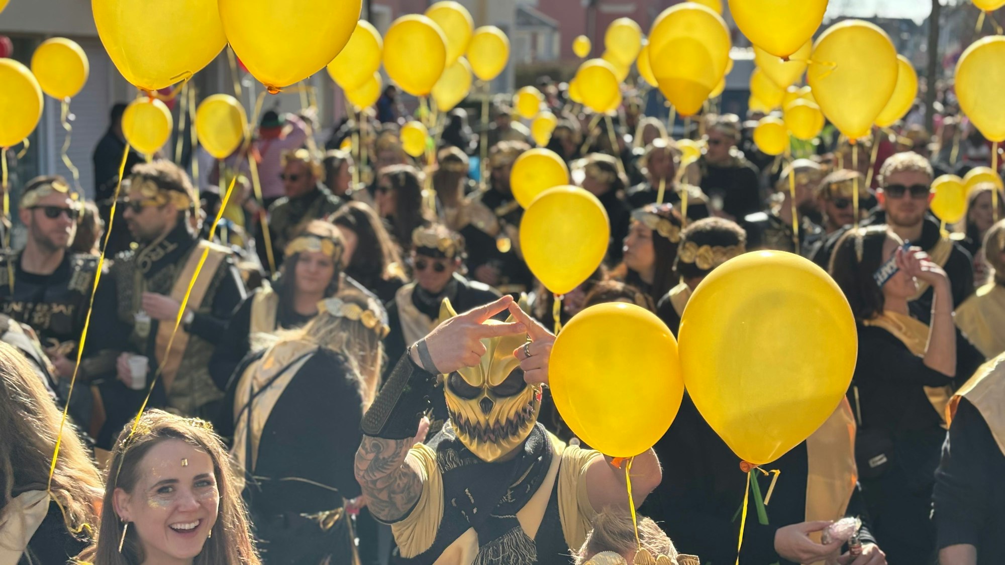 Menschen in schwarz-goldenen Kostümen tragen gelbe Luftballons. Ein maskierter Mann