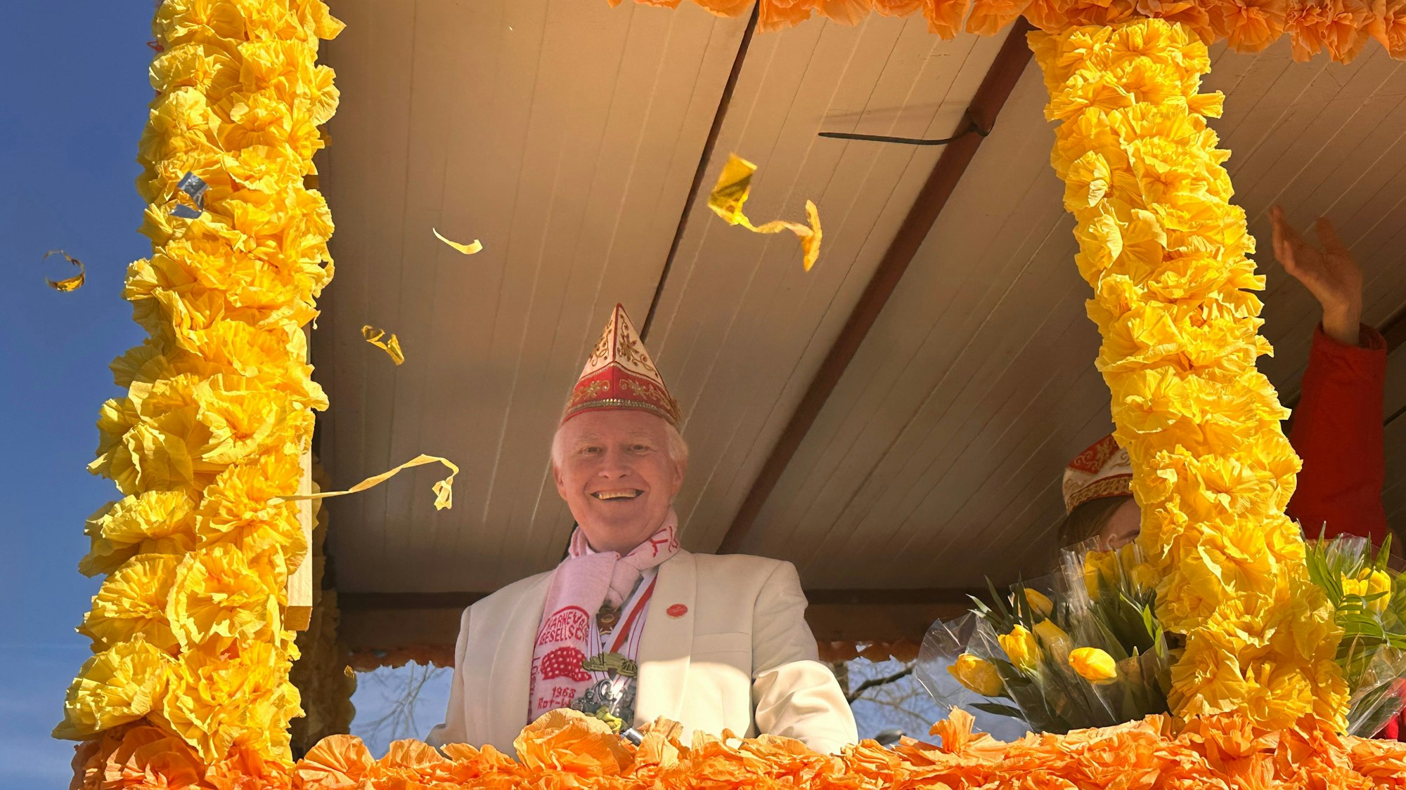 Hinter einem Rahmen aus Papierblumen ist ein Mann mit Narrenkappe zu sehen.