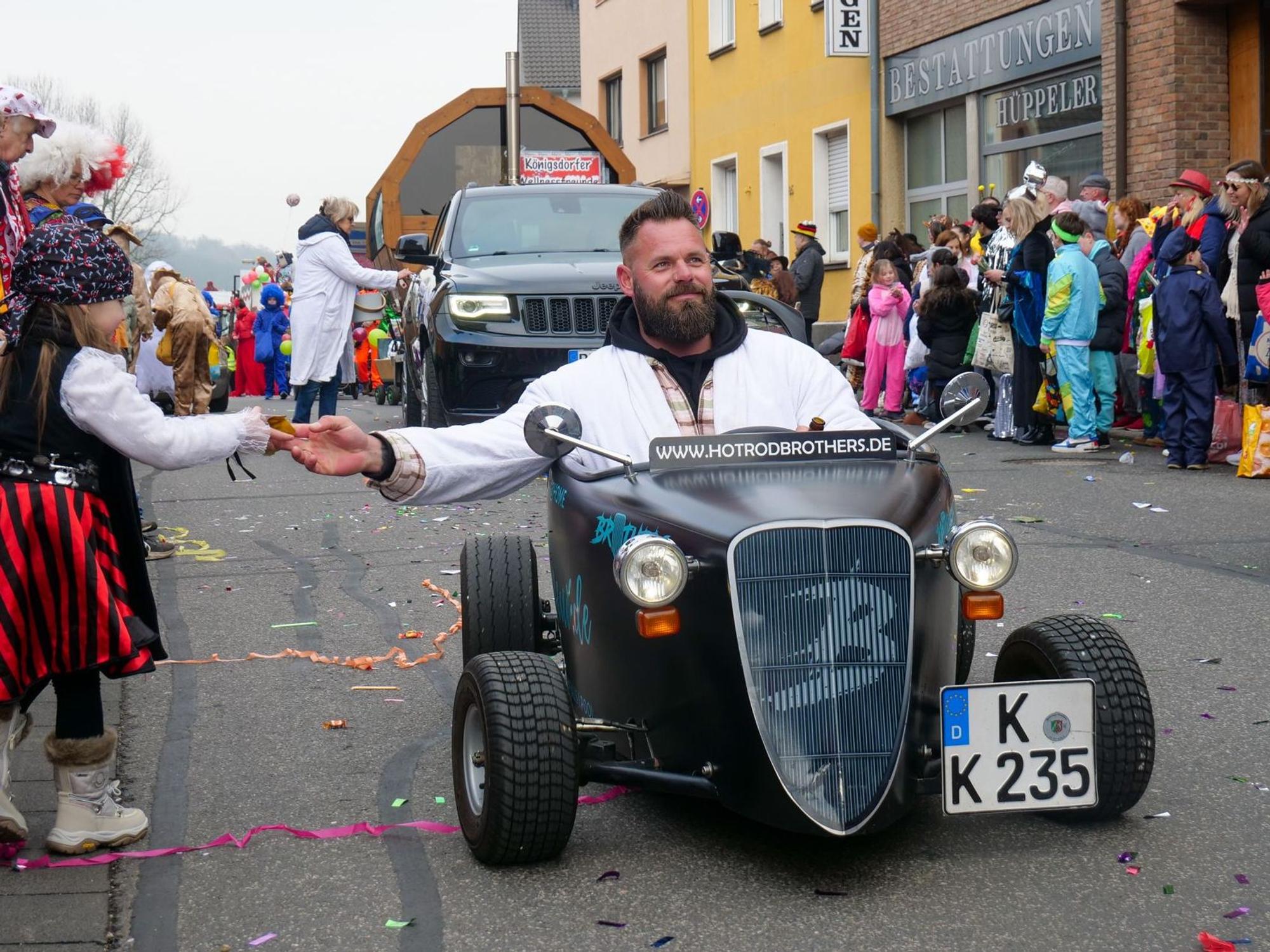 Auf dem Bild ist ein Mann in einem Miniauto zu sehen, der einem Mädchen am Zugweg Kamelle zu steckt.