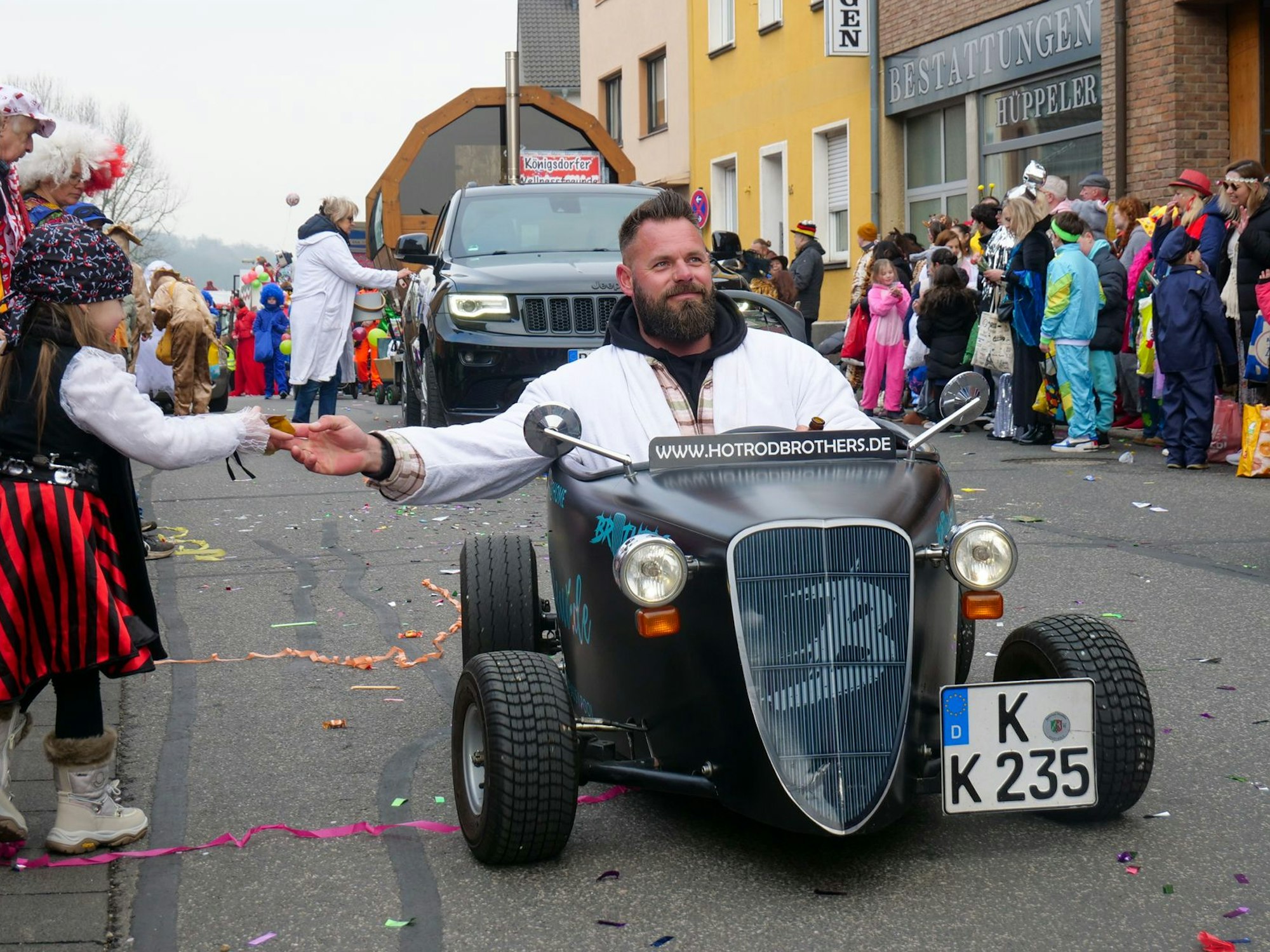 Auf dem Bild ist ein Mann in einem Miniauto zu sehen, der einem Mädchen am Zugweg Kamelle zu steckt.