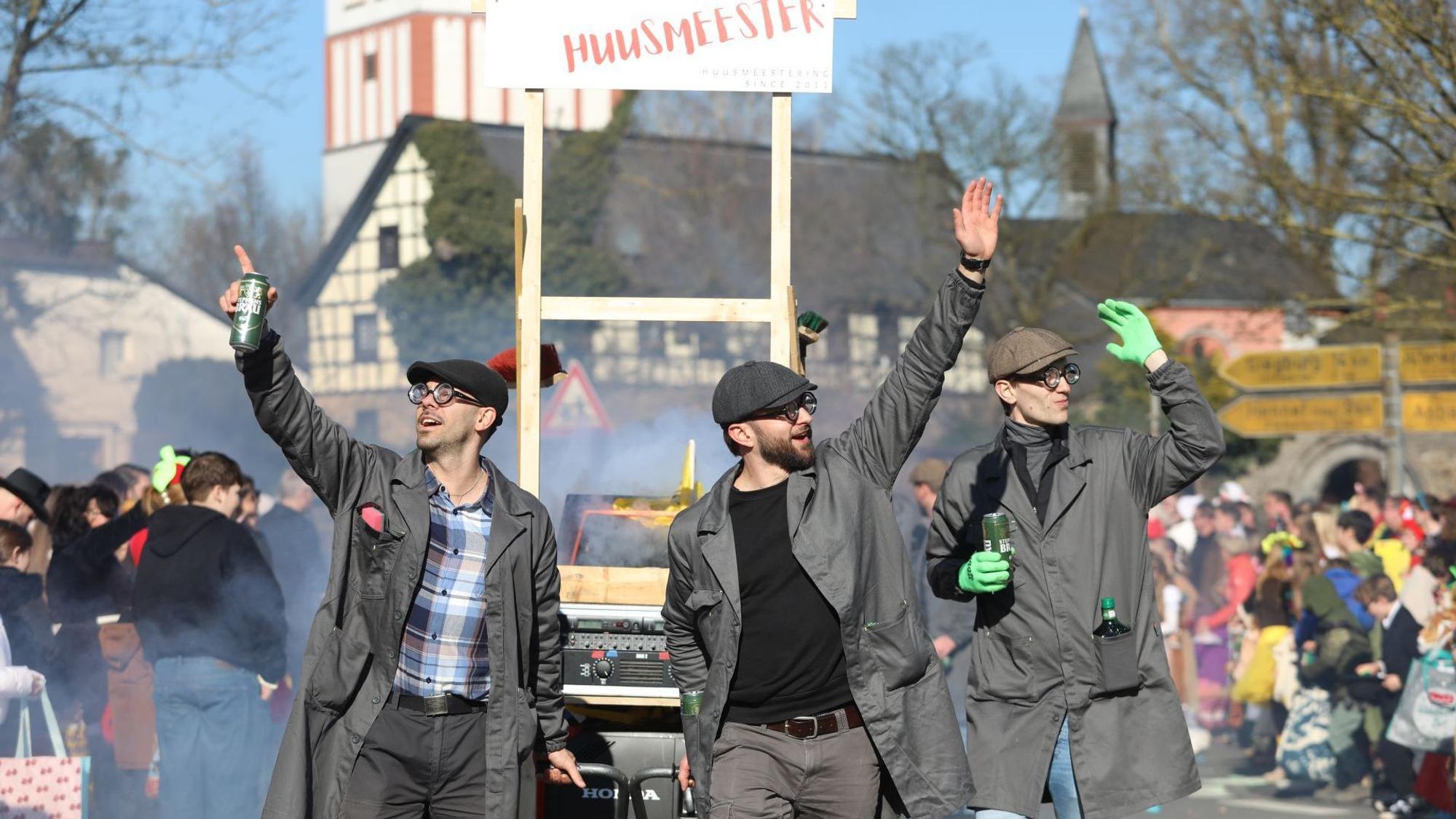 Drei Männer in Hausmeisterkleidung ziehen einen Karnevalswagen, im Hintergrund die Oberpleiser Pfarrkirche.
