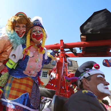 Drei bunt kostümierte und geschminkte Frauen als Clowns auf einem Karnevalswagen.