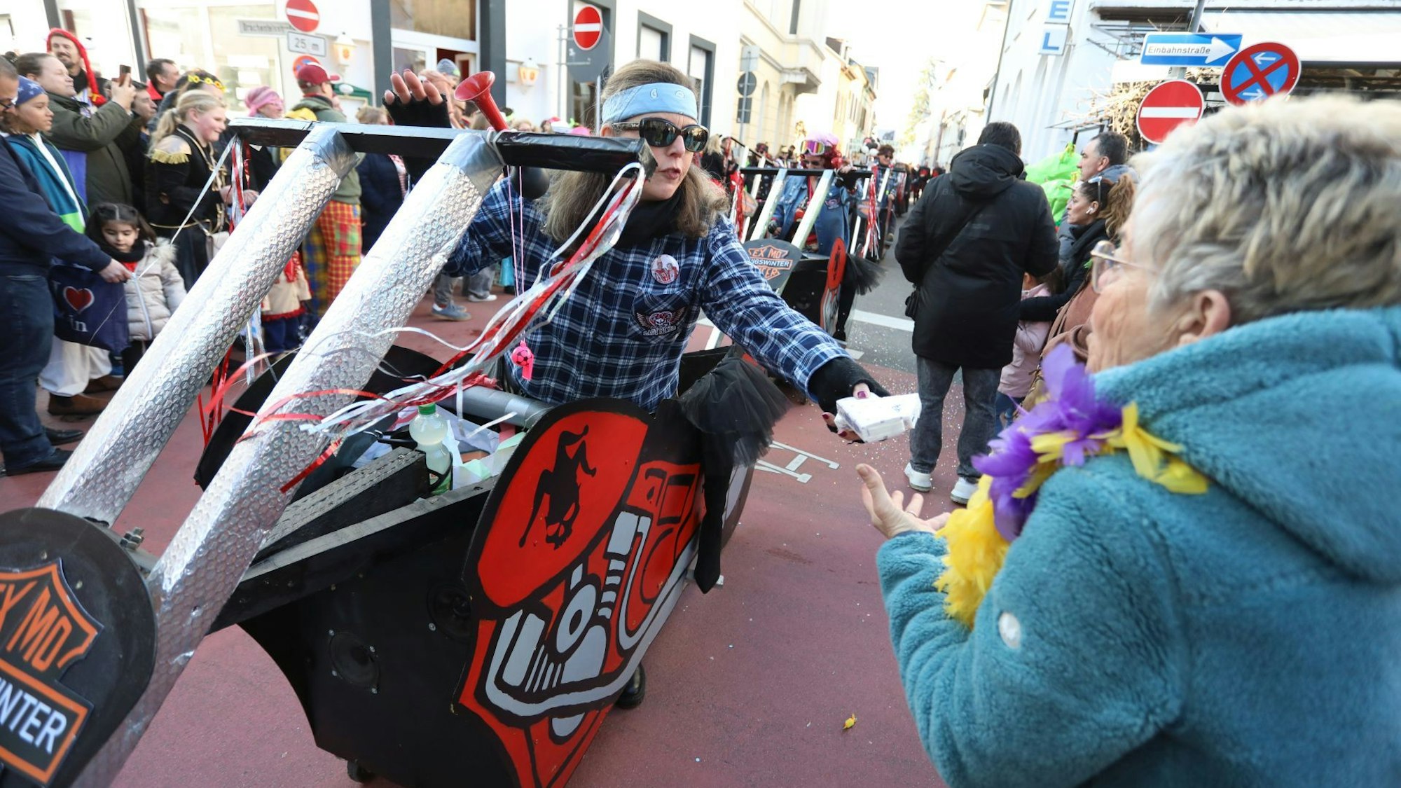 Mit Motorrädern aus Pappe verteilen verkleidete Jecken Kamelle an Zuschauer am Straßenrand.