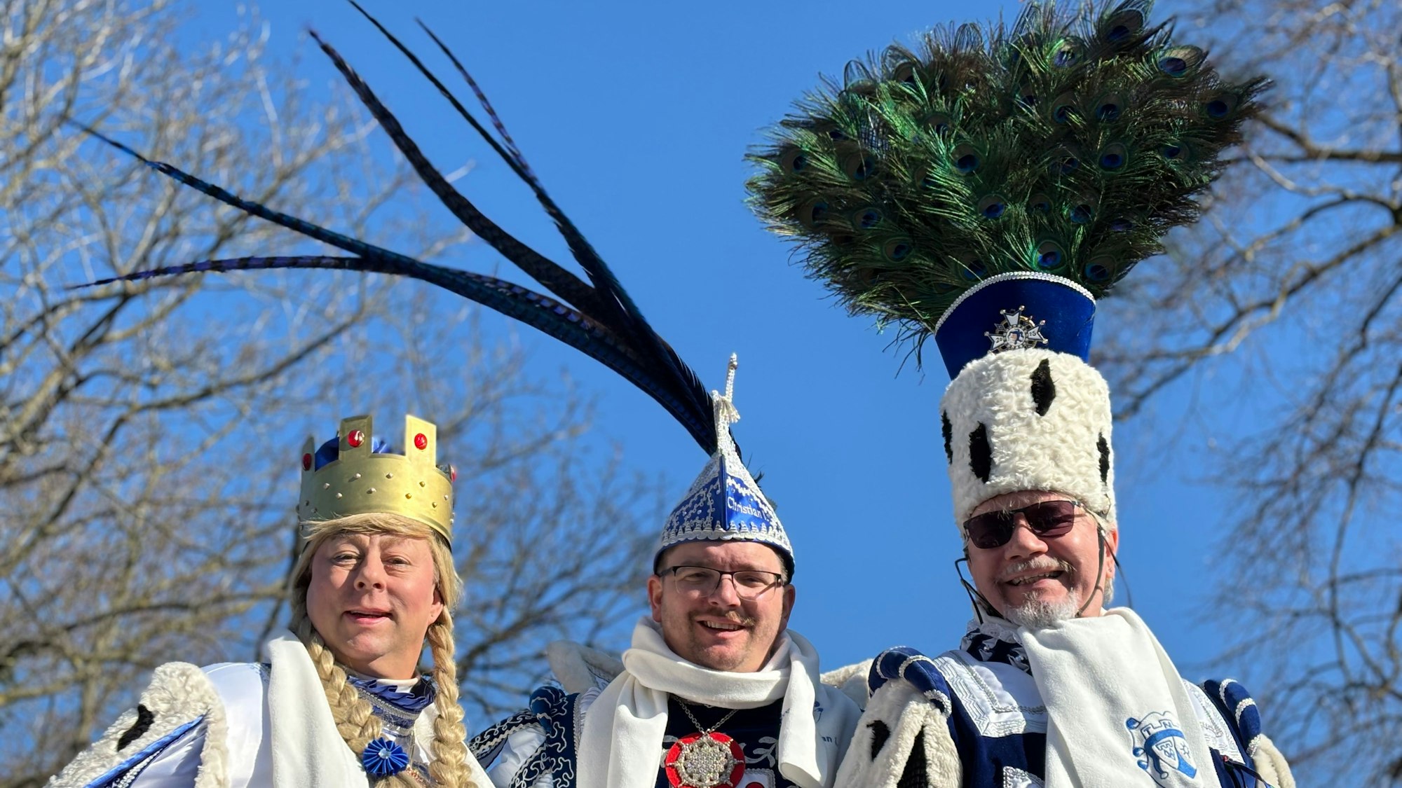 Ein karnevalistisches Dreigestirn schaut vor einem blauen Himmel mit Bäumen im Hintergrund in die Kamera.