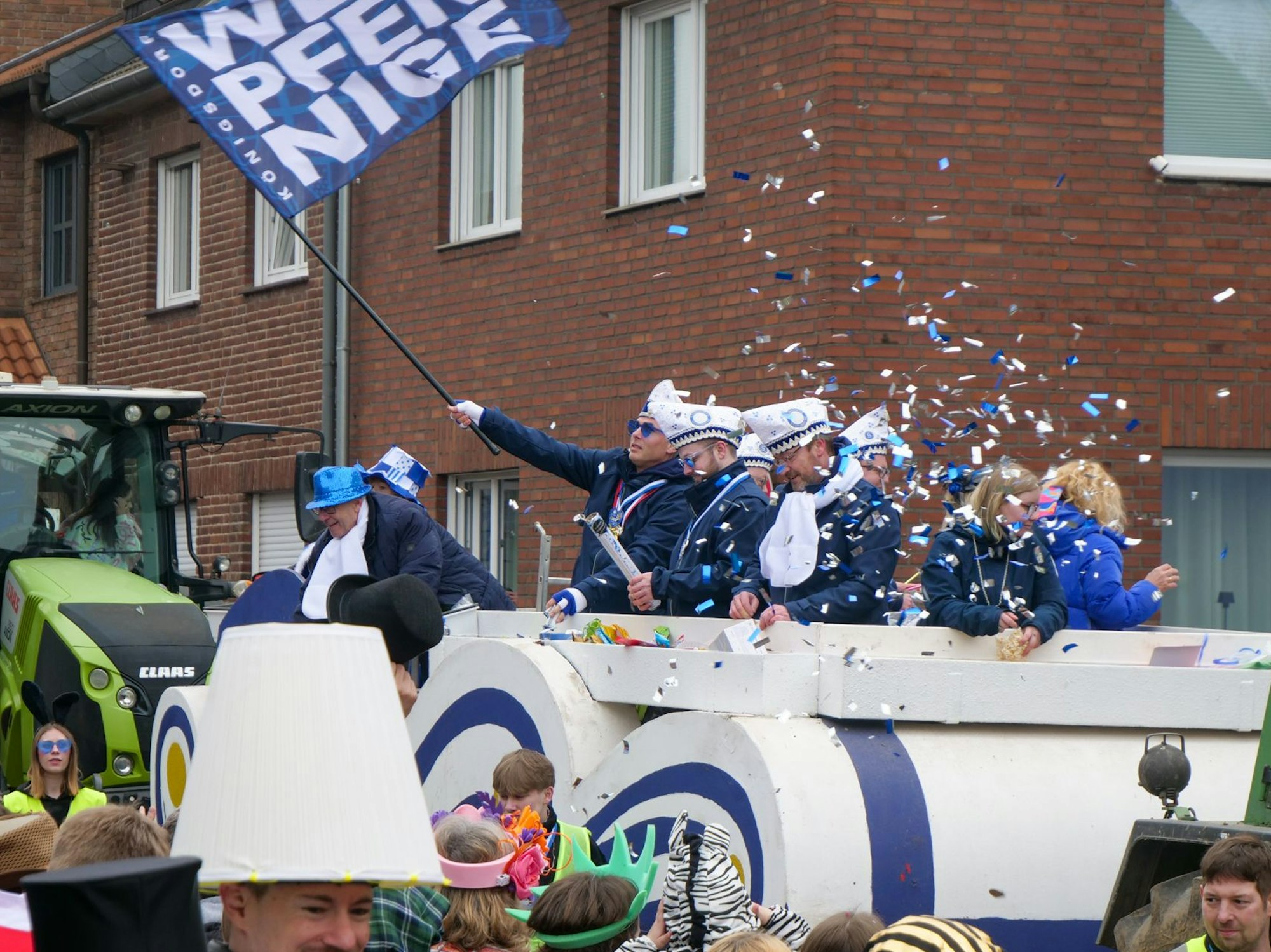 Auf dem Bild ist ein Festwagen der KG Königsdorfer Weißpfennige zu sehen, ein blau-weißer Konfettiregen wird von den Teilnehmern geworfen.