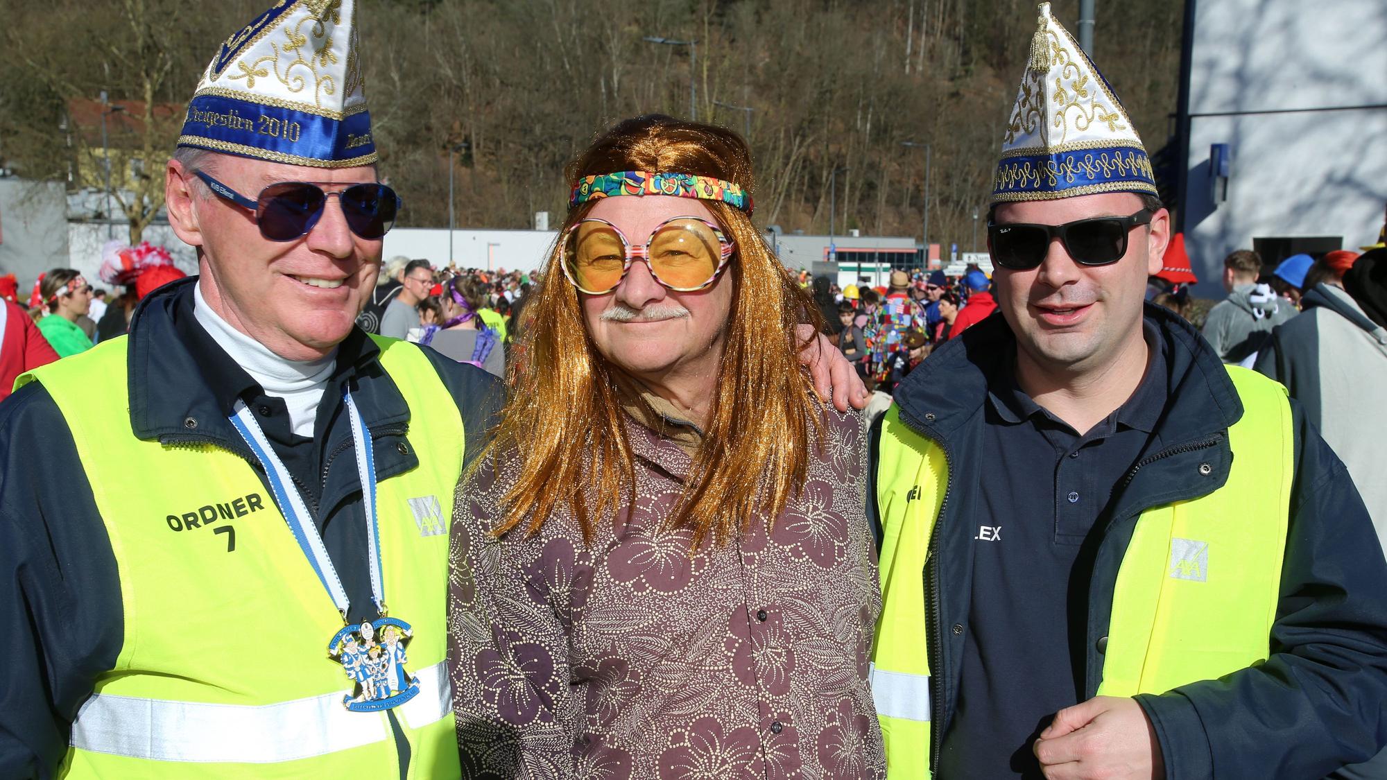 Ein als Hippie verkleideter Mann und zwei Ordner bei einem Karnevalszug.
