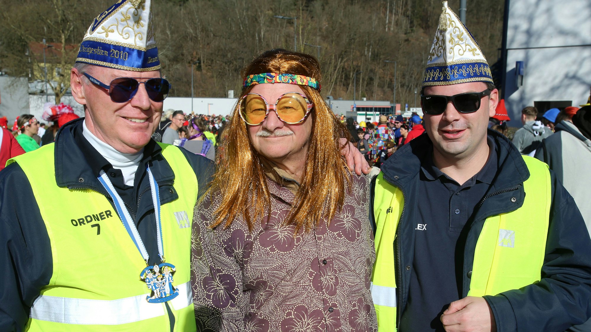 Ein als Hippie verkleideter Mann und zwei Ordner bei einem Karnevalszug.
