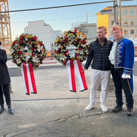 Am Ort der Katastrophe: Christoph Kuckelkorn, Marvin Pagel, der Bruder eines der Opfer, Henriette Reker, Trompeter der Roten Funken (v.r.) Die vier Menschen stehen an einem Gitterzaun vor der Baustelle; an dem Zaun hängen zwei Kränze für die beiden Opfer mit Blumenschmuck in rot, weiß, orange und hellgrün sowie rot-weißen Gedenkschleifen.