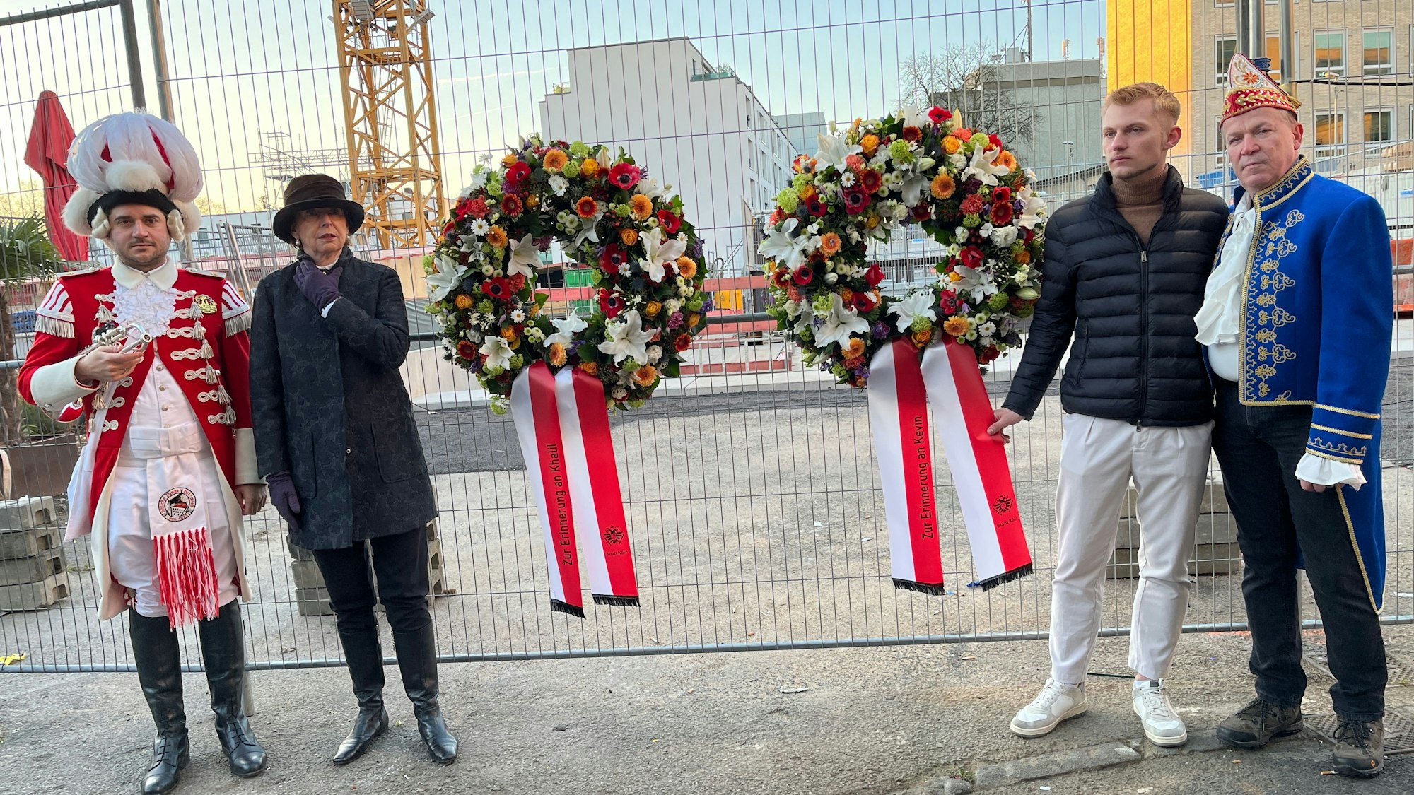 Am Ort der Katastrophe: Christoph Kuckelkorn, Marvin Pagel, der Bruder eines der Opfer, Henriette Reker, Trompeter der Roten Funken (v.r.) Die vier Menschen stehen an einem Gitterzaun vor der Baustelle; an dem Zaun hängen zwei Kränze für die beiden Opfer mit Blumenschmuck in rot, weiß, orange und hellgrün sowie rot-weißen Gedenkschleifen.